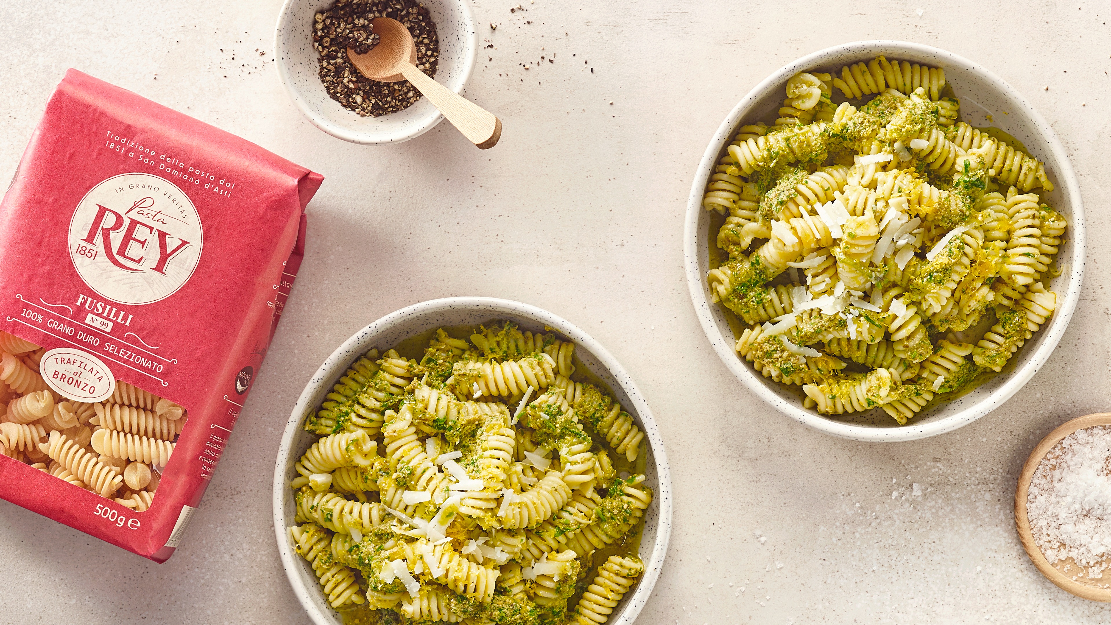 Zwei Schalen mit gedrehten Nudeln, grüner Soße und geriebenem Käse. Daneben eine Packung Pasta Rey Fusilli.