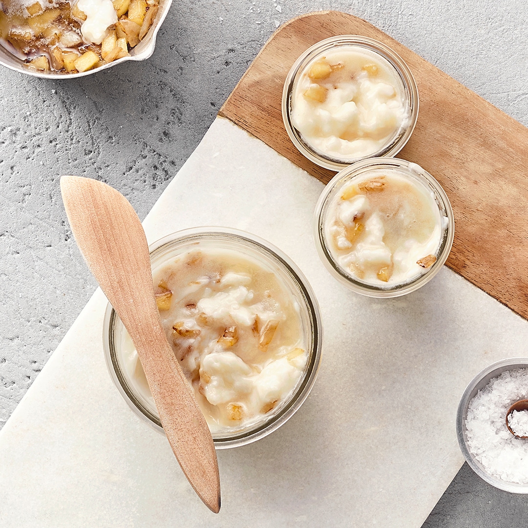 Drei Glasschalen mit Gänseschmalz und Apfelstücken stehen auf einem Brett. Oben links befindet sich eine Schüssel mit Äpfeln, unten rechts eine Schüssel mit Salz.