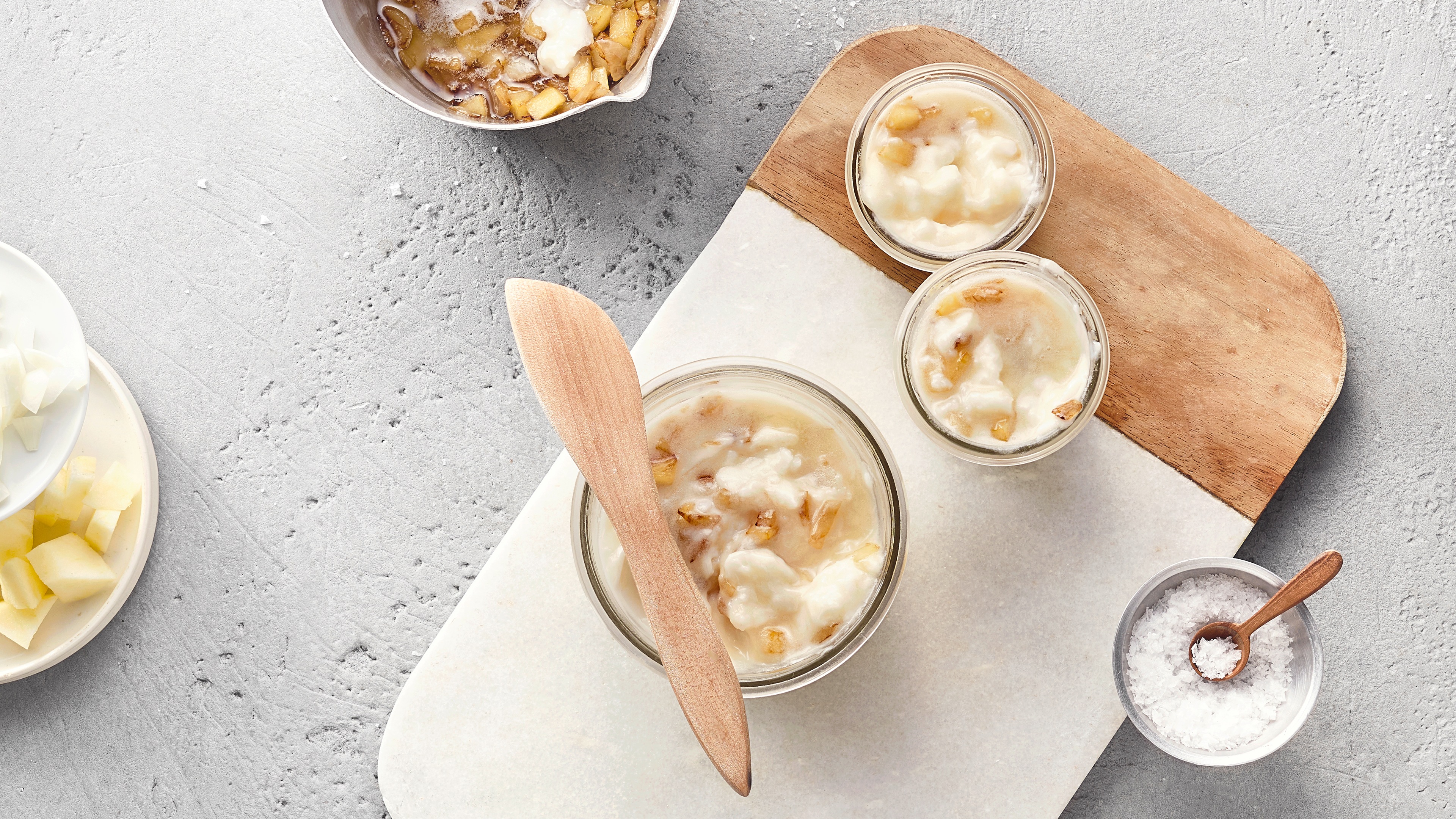 Drei Glasschalen mit Gänseschmalz und Apfelstücken stehen auf einem Brett. Oben links befinden sich Schüsseln mit Äpfeln, unten rechts eine Schüssel mit Salz.