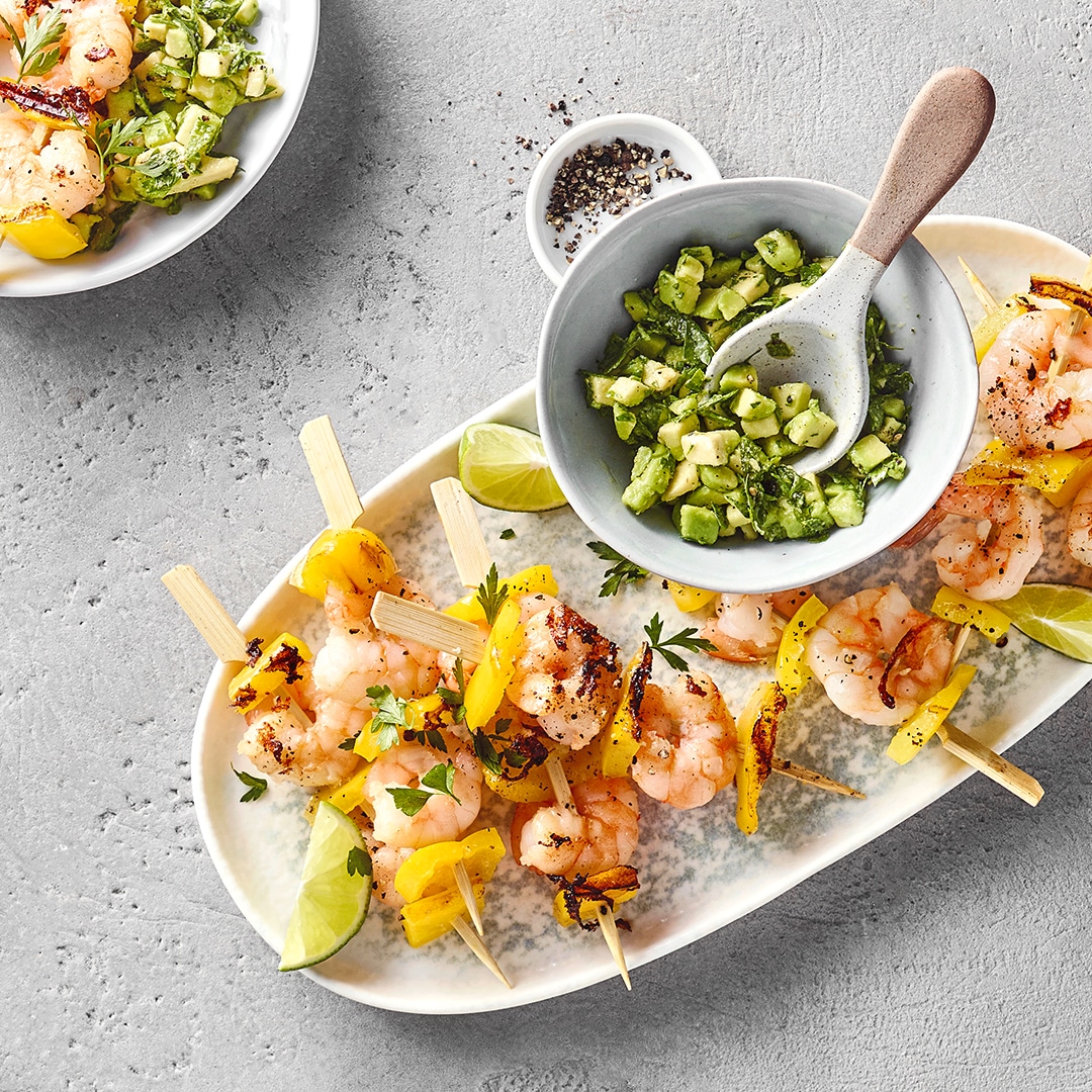 Gegrillte Garnelenspieße mit gelber Paprika und Limettenspalten, serviert mit Avocado-Salsa.