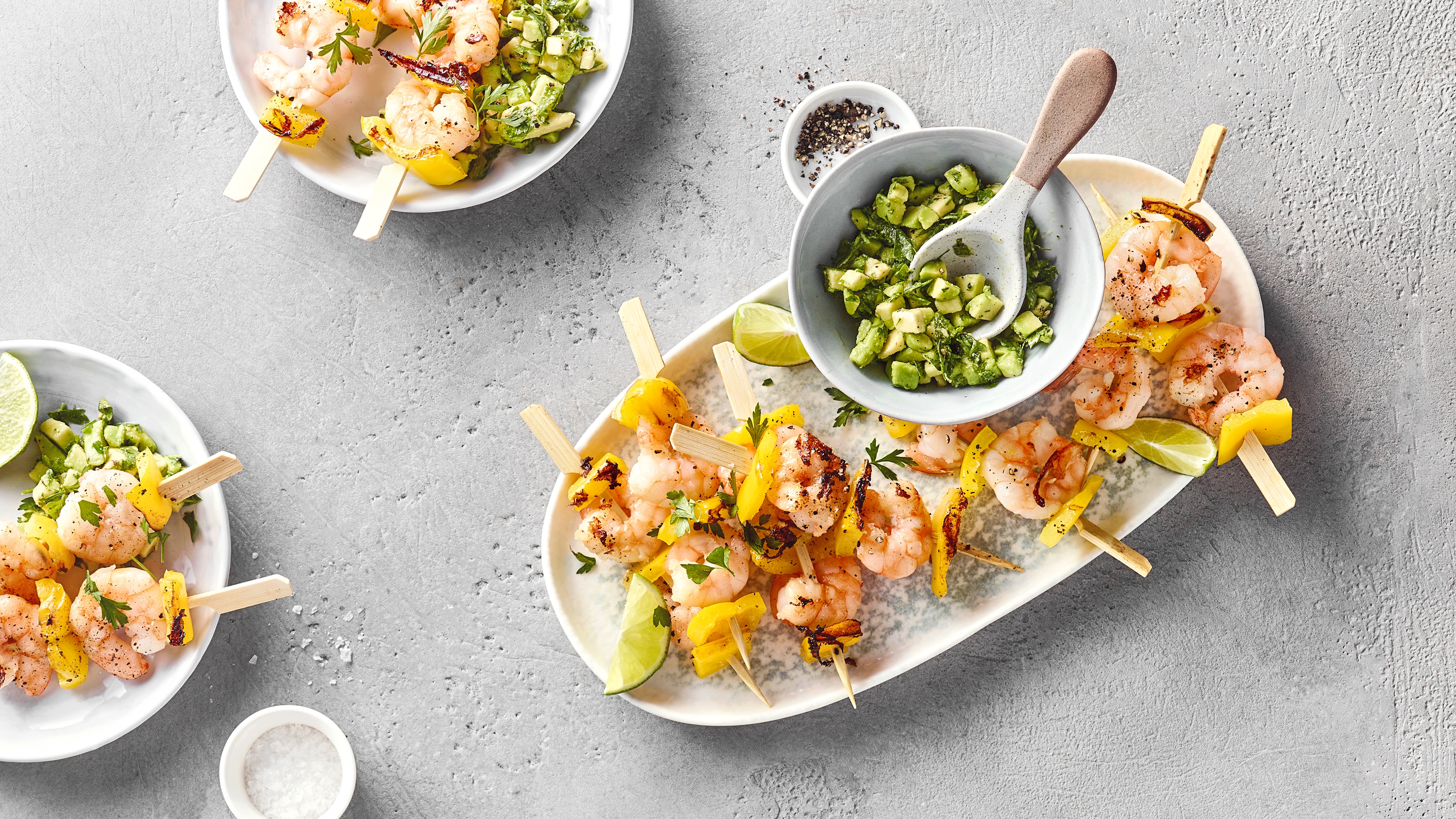 Gegrillte Garnelenspieße mit gelber Paprika und Limettenspalten, serviert mit Avocado-Salsa.