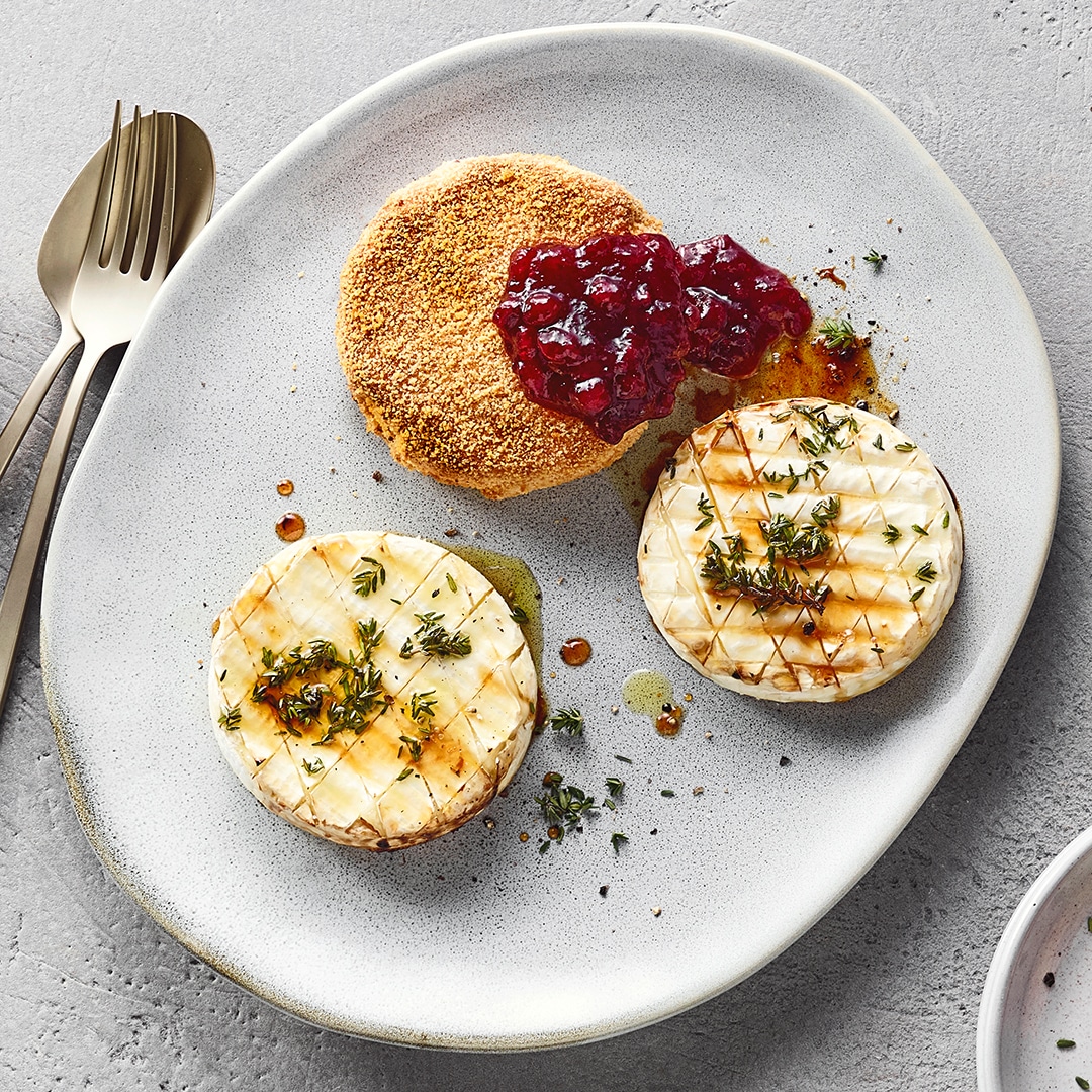 Ein Teller mit zwei runden Camemberts mit Kräutern und heller Soße sowie einem panierten Käsetaler mit roter Beerensoße.