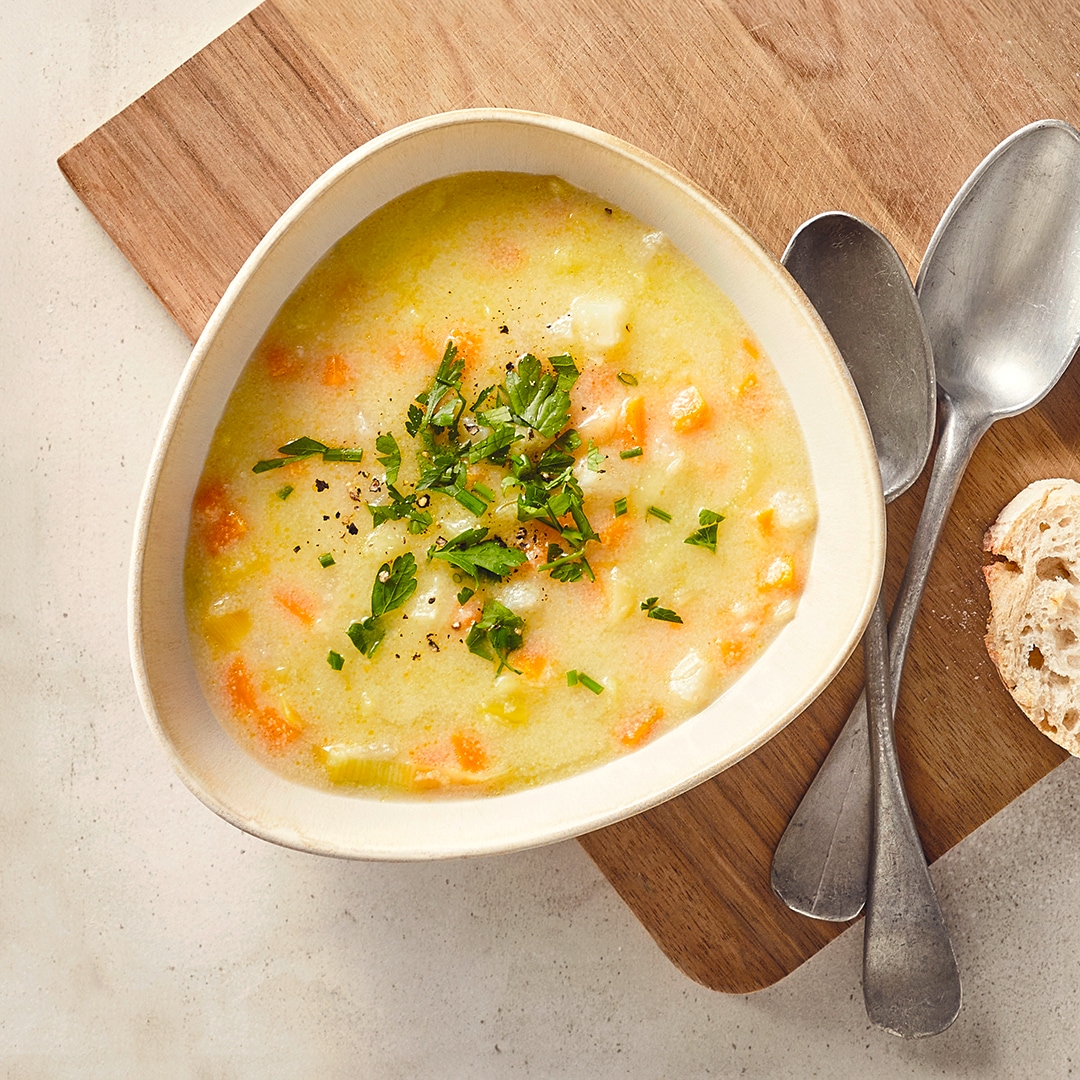 Eine Schale mit einer hellen Suppe, mit Karottenstücken und Lauch, garniert mit gehackter Petersilie, daneben ein Stück Brot