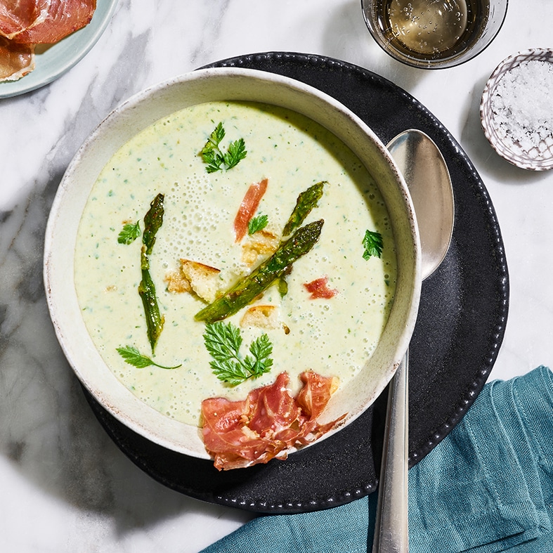 Schale mit cremiger Suppe, garniert mit Spargelstücken, knusprigen Schinken‑Chips und Petersilie. Rechts daneben liegt ein Löffel. Oben links befinden sich weitere Schinken‑Chips, oben rechts steht eine Schale mit Salz.