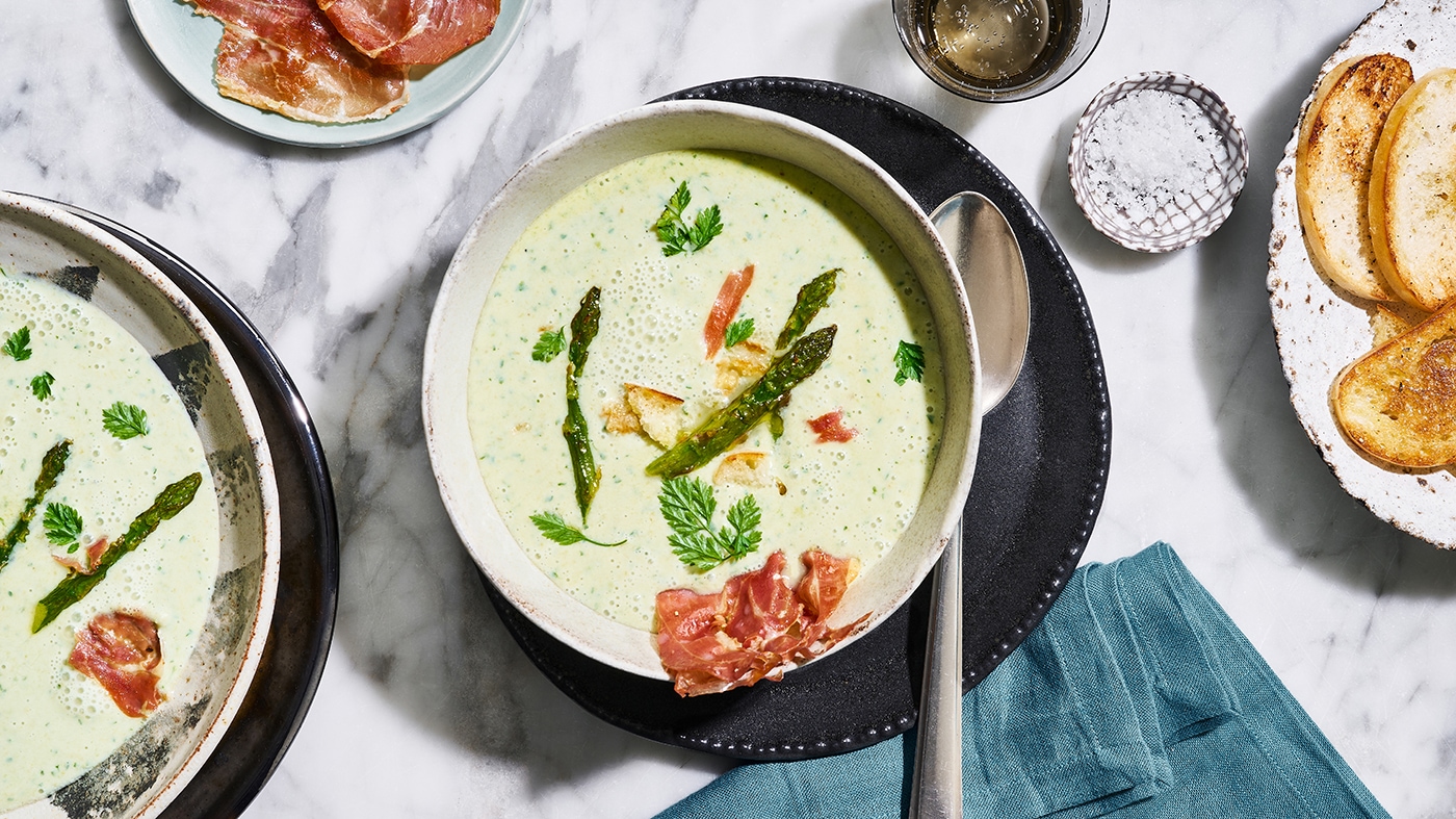 Schale mit cremiger Suppe, garniert mit Spargelstücken, knusprigen Schinken‑Chips und Petersilie. Rechts daneben liegt ein Löffel. Oben links befinden sich weitere Schinken‑Chips, oben rechts steht ein Salzbehälter. Links neben der Hauptschale ist ein weiterer Suppenteller teilweise zu sehen. Rechts daneben liegen mehrere knusprige Brotscheiben.