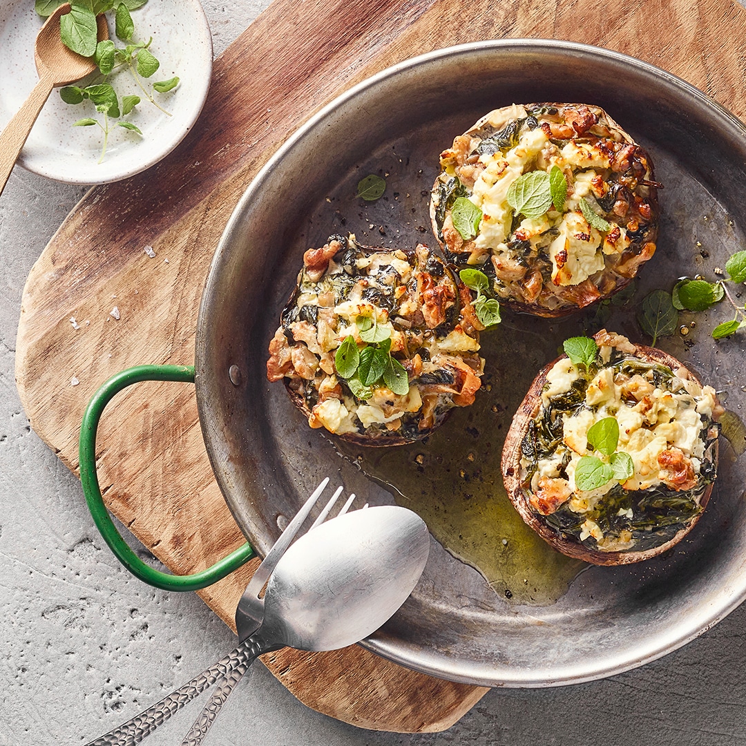 Eine Pfanne mit drei gefüllten braunen Pilzkappen, belegt mit weißer & grüner Mischung sowie kleinen grünen Blättern & Öl.