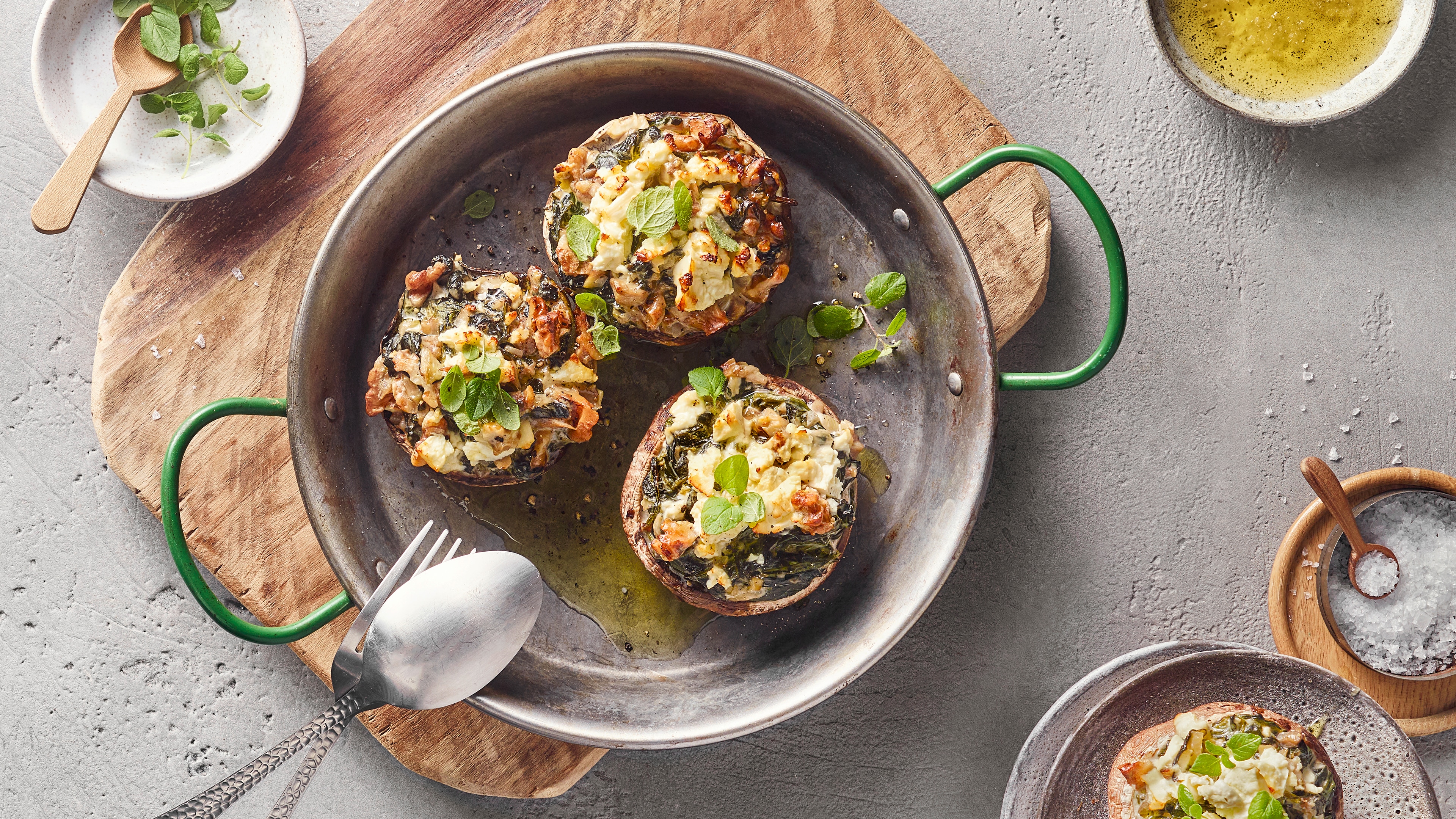 Eine Pfanne mit drei gefüllten braunen Pilzkappen, belegt mit weißer & grüner Mischung sowie kleinen grünen Blättern & Öl.