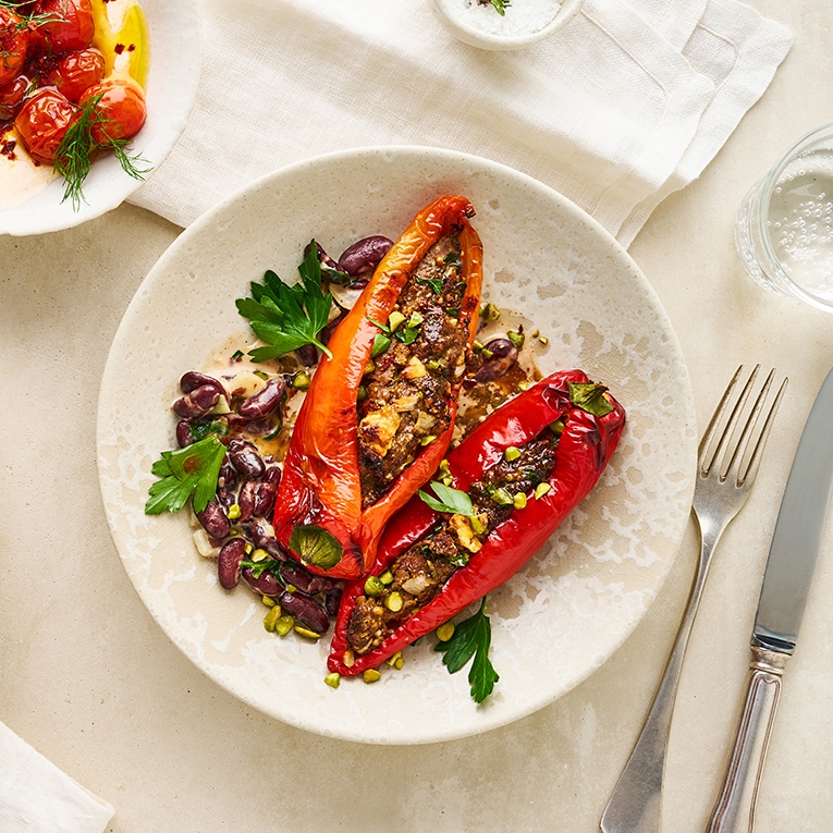 Rote Spitzpaprika, gefüllt mit Hackfleisch und Feta, in heller Soße mit Kidneybohnen, getoppt mit Pistazien, daneben eine Schale mit Dip und geschmorte Tomaten