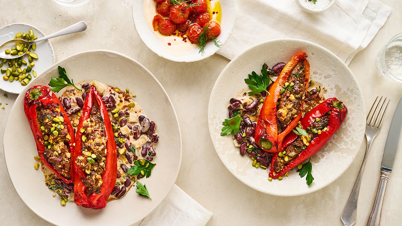 Rote Spitzpaprika, gefüllt mit Hackfleisch und Feta, in heller Soße mit Kidneybohnen, getoppt mit Pistazien, daneben eine Schale mit Dip und geschmorte Tomaten