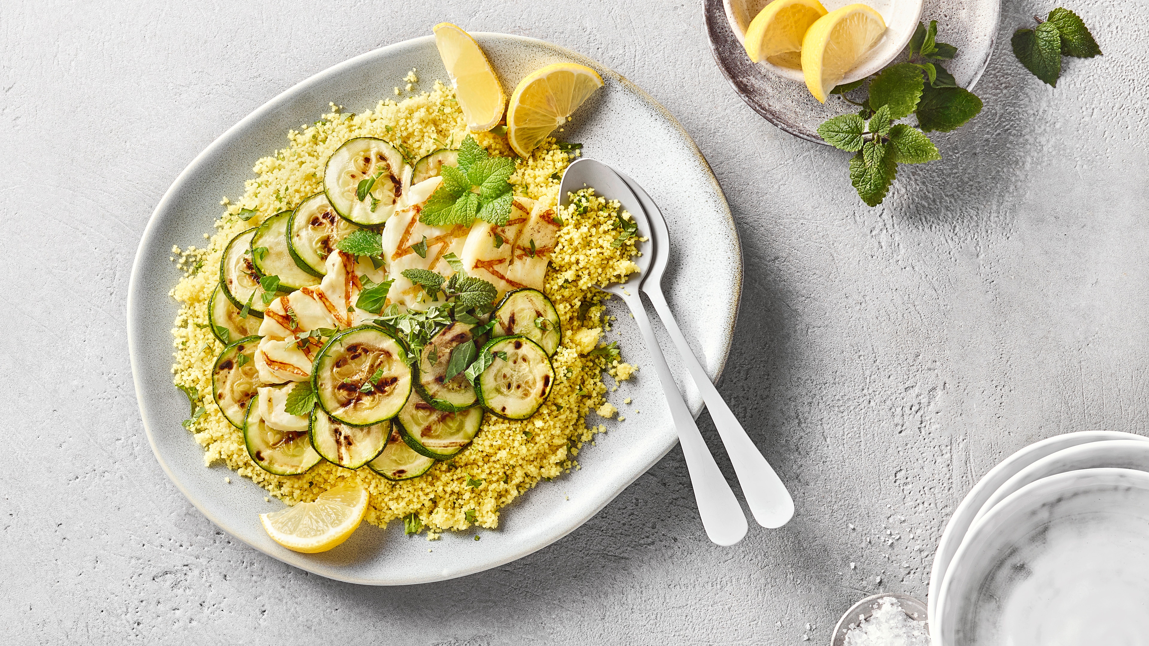 Großer Teller Couscous mit gegrillter Zucchini und Halloumi, garniert mit Minze und Zitronenspalten.