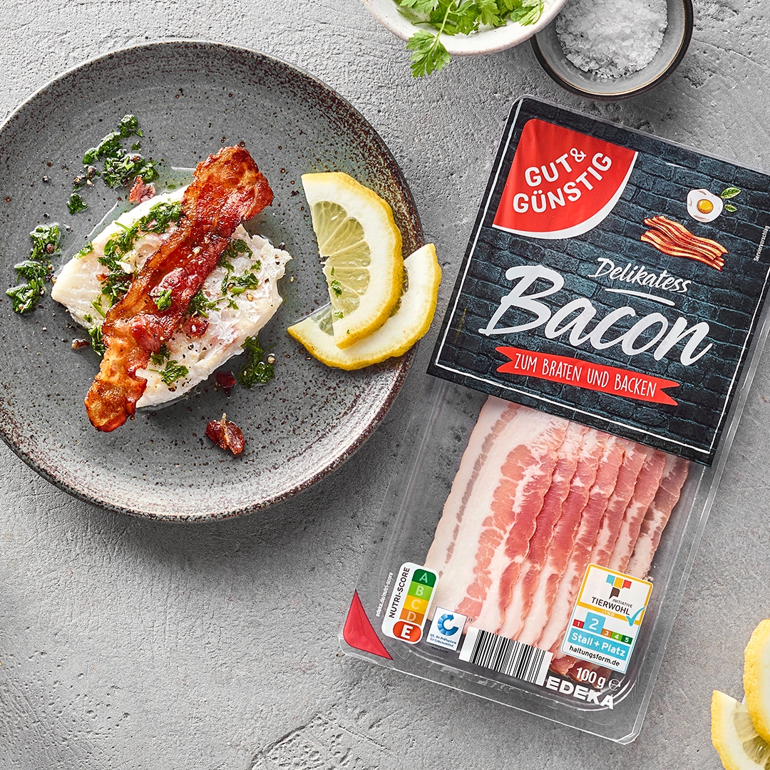 Teller mit Fischfilet, Speckscheibe und Zitronenscheiben. Daneben eine Packung Gut&Günstig Bacon.