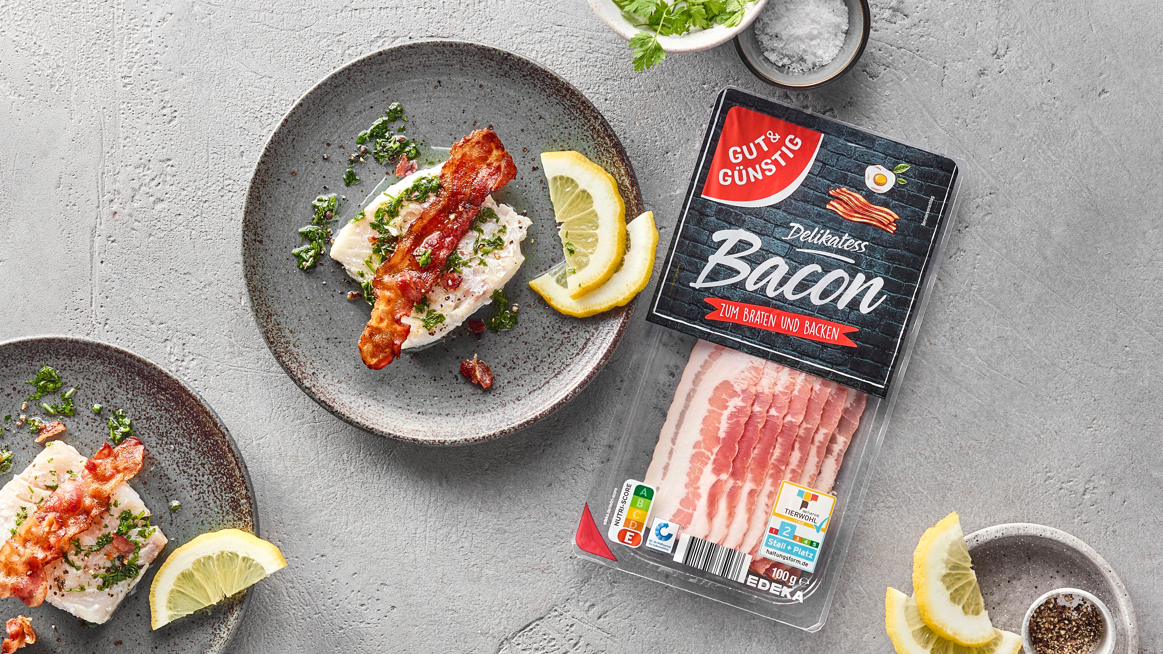 Zwei Teller mit Fischfilet, Speckscheibe und Zitronenscheiben. Daneben eine Packung Gut&Günstig Bacon.
