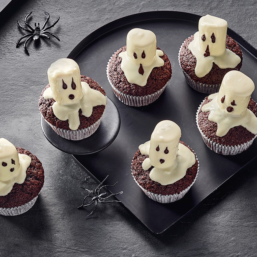 Dunkle Muffins mit kleinen weißen Geister-Dekorationen aus Glasur auf einem dunklen Tablett, umgeben von Halloween-Spinnendeko.