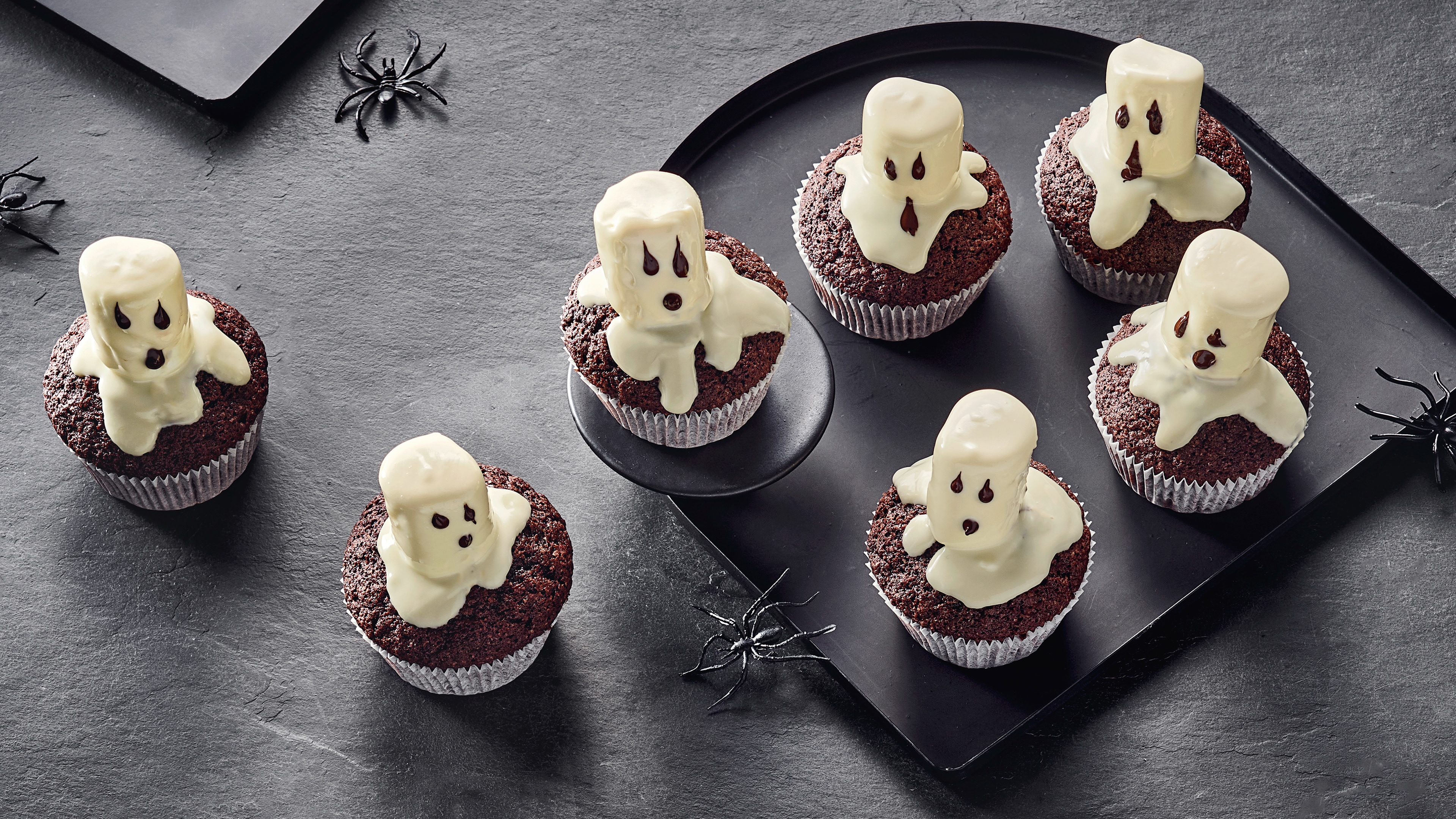 Schokoladen-Cupcakes mit kleinen weißen Geister-Dekorationen aus Glasur auf einem dunklen Tablett, umgeben von Halloween-Spinnendeko.