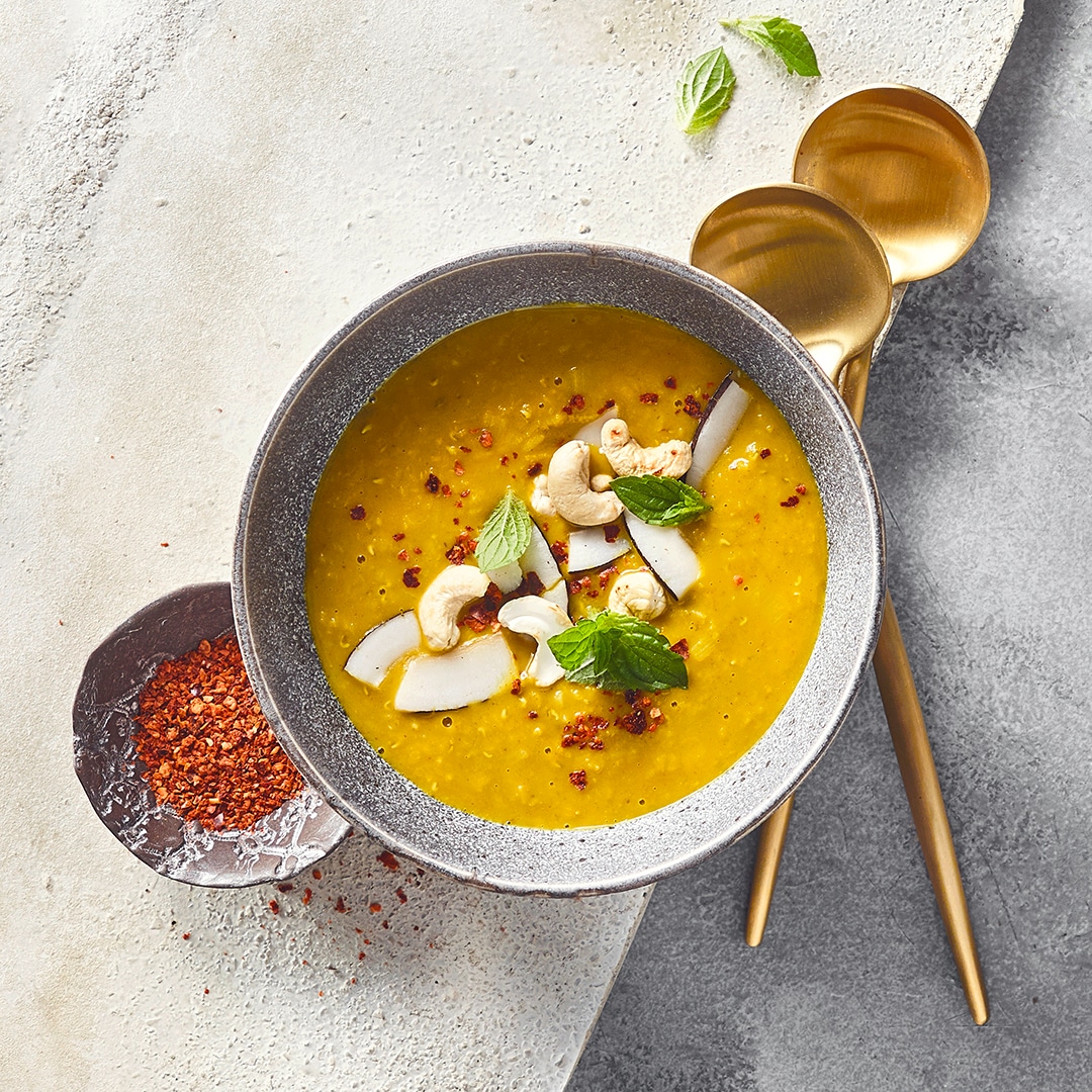 Ein Schälchen mit curryfarbener Suppe,  garniert mit Kokosraspeln, Cashewkernen und frischen Minzblättern