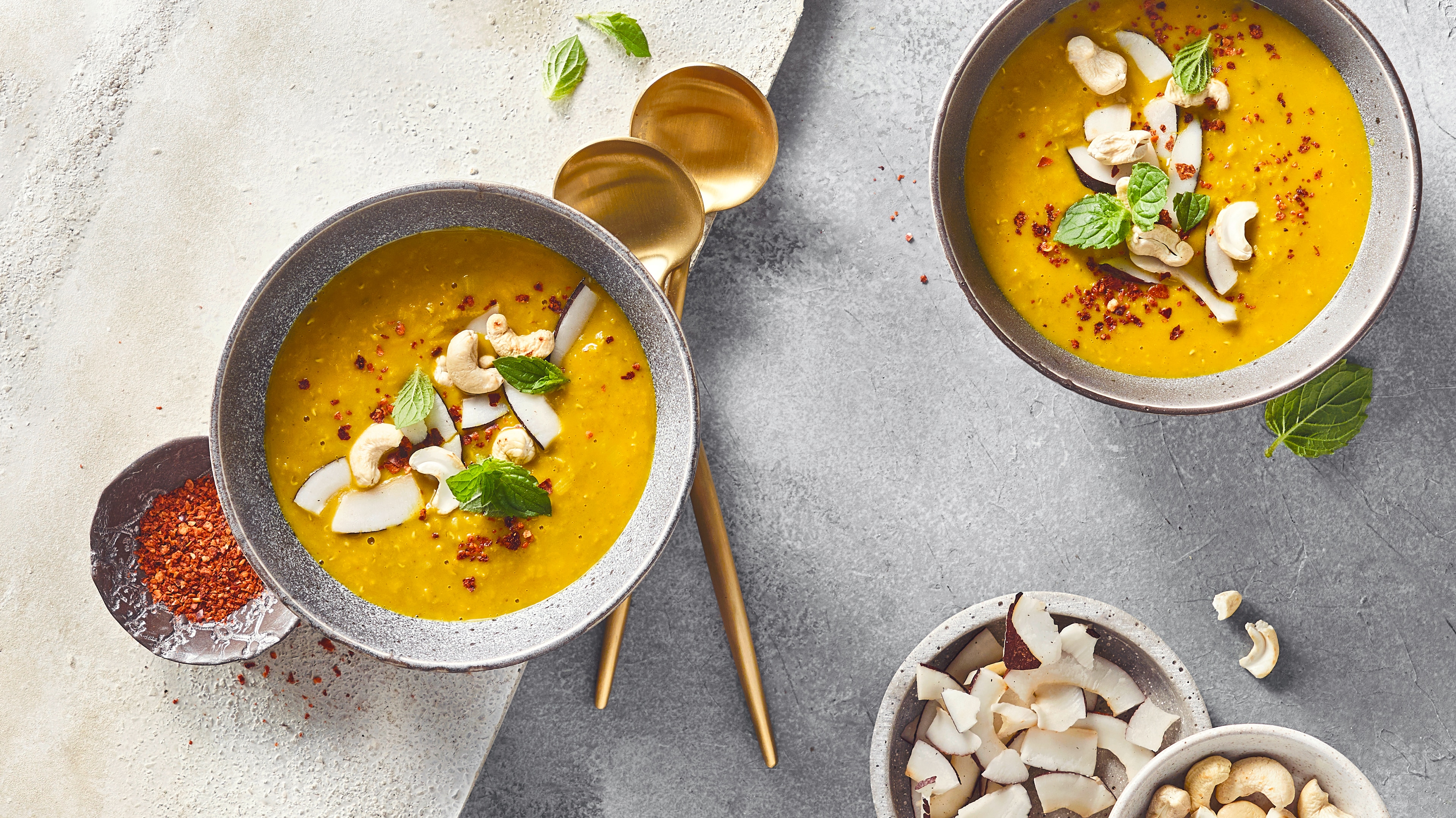 Ein Schälchen mit curryfarbener Suppe,  garniert mit Kokosraspeln, Cashewkernen und frischen Minzblättern