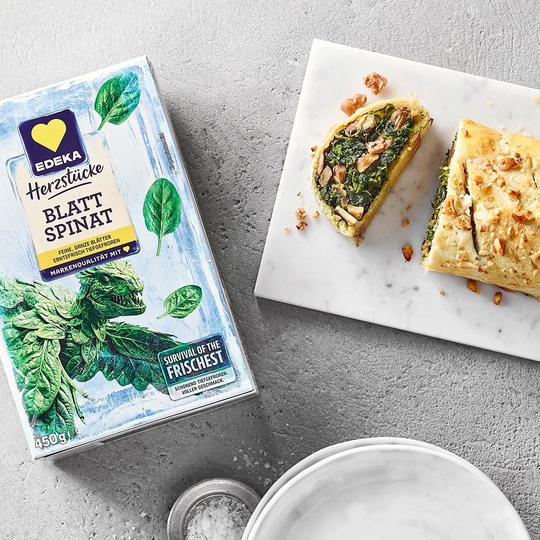 Länglicher Strudel mit Spinat und Nüssen gefüllt auf Marmorplatte. Daneben eine Packung EDEKA Herzstücke Blattspinat.
