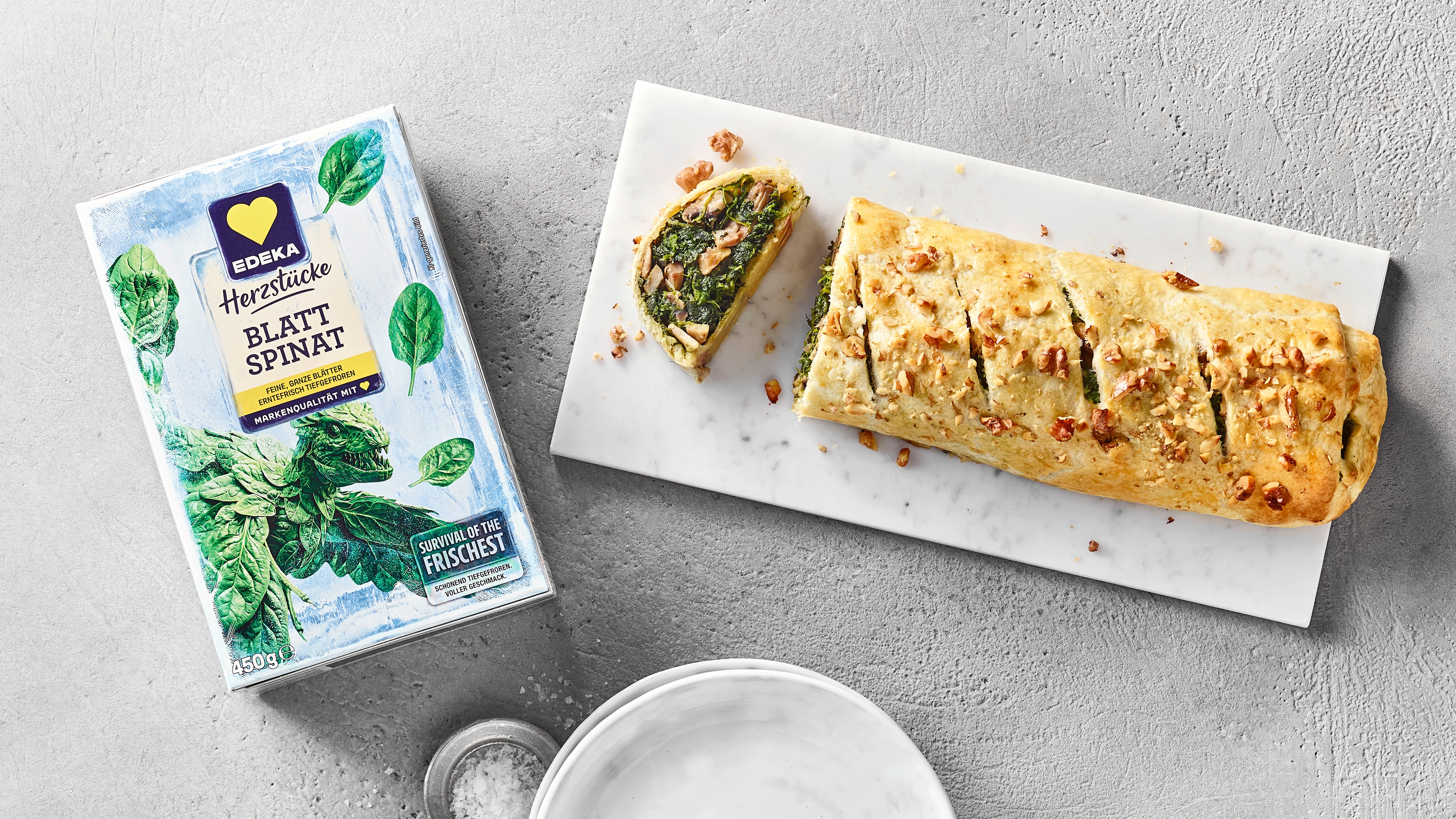 Länglicher Strudel mit Spinat und Nüssen gefüllt auf Marmorplatte. Daneben eine Packung EDEKA Herzstücke Blattspinat.