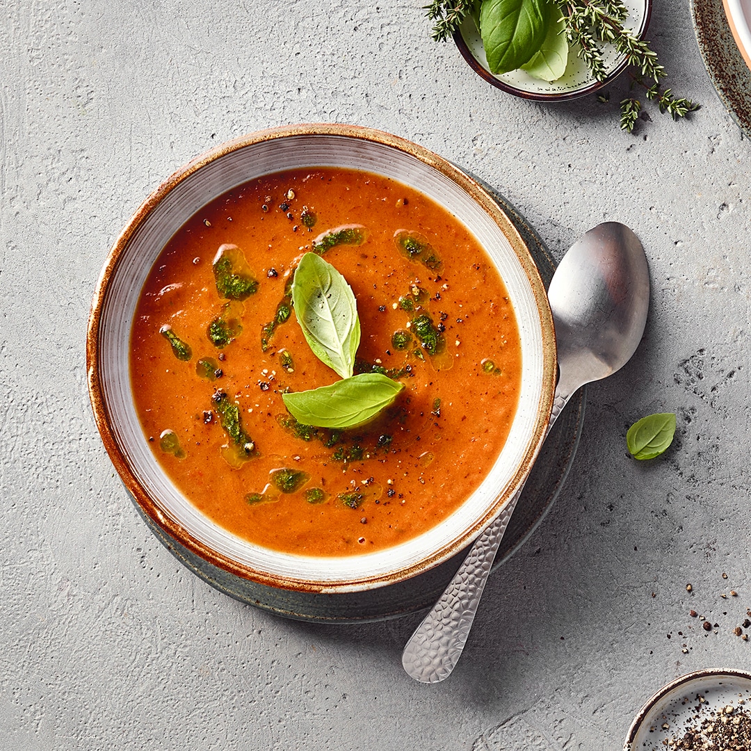Eine Schüssel mit roter pürierter Suppe, darauf frische Kräuterblätter und grüne Öltropfen.