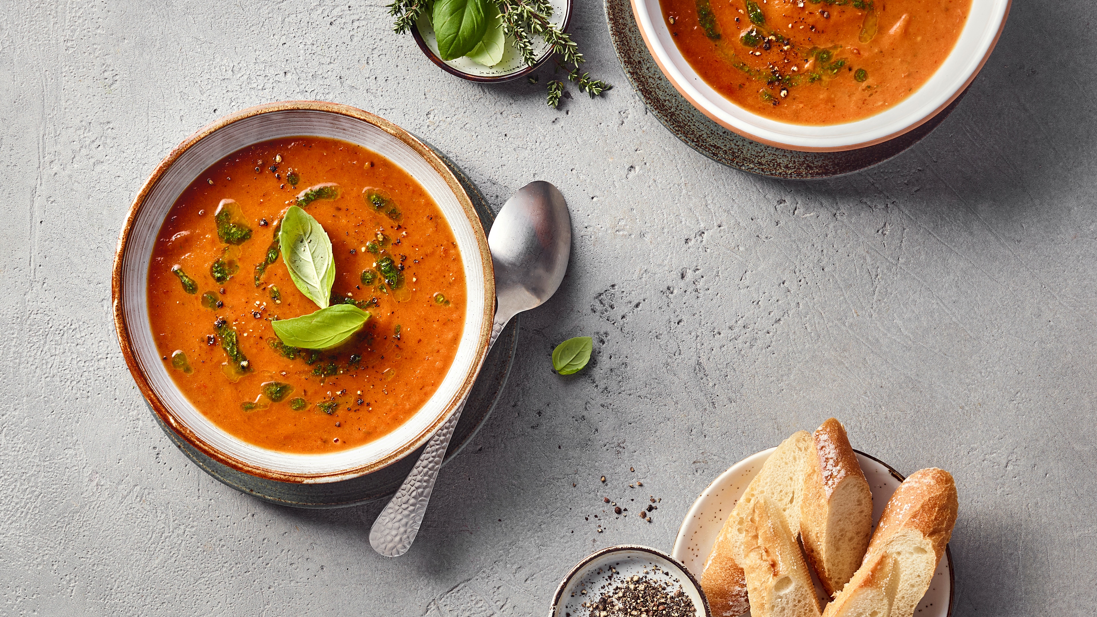 Eine Schüssel mit roter pürierter Suppe, darauf frische Kräuterblätter und grüne Öltropfen.