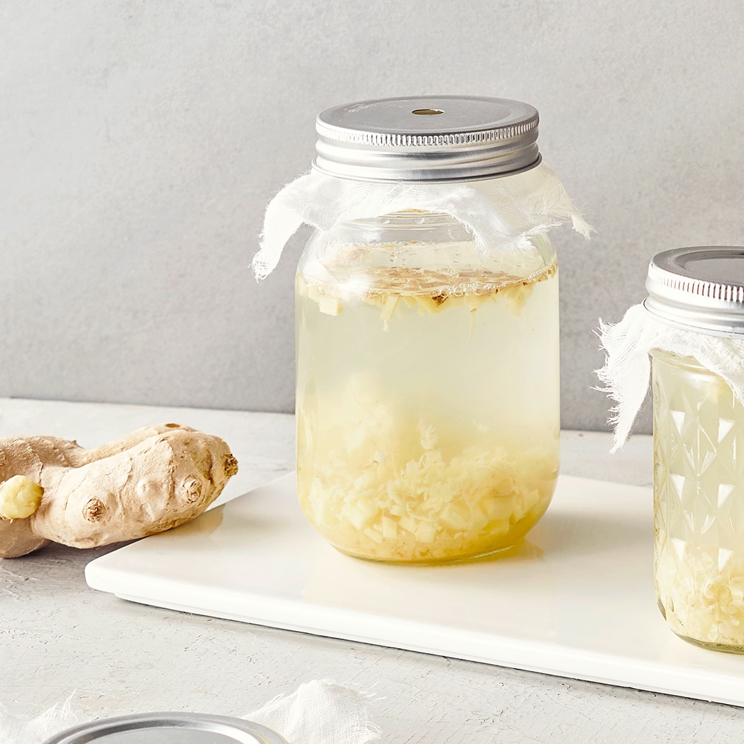 Ein Glas mit heller Flüssigkeit und feinen, geriebenen Stücken am Boden, daneben ein Stück frischer Ingwer auf einem weißen Brett.