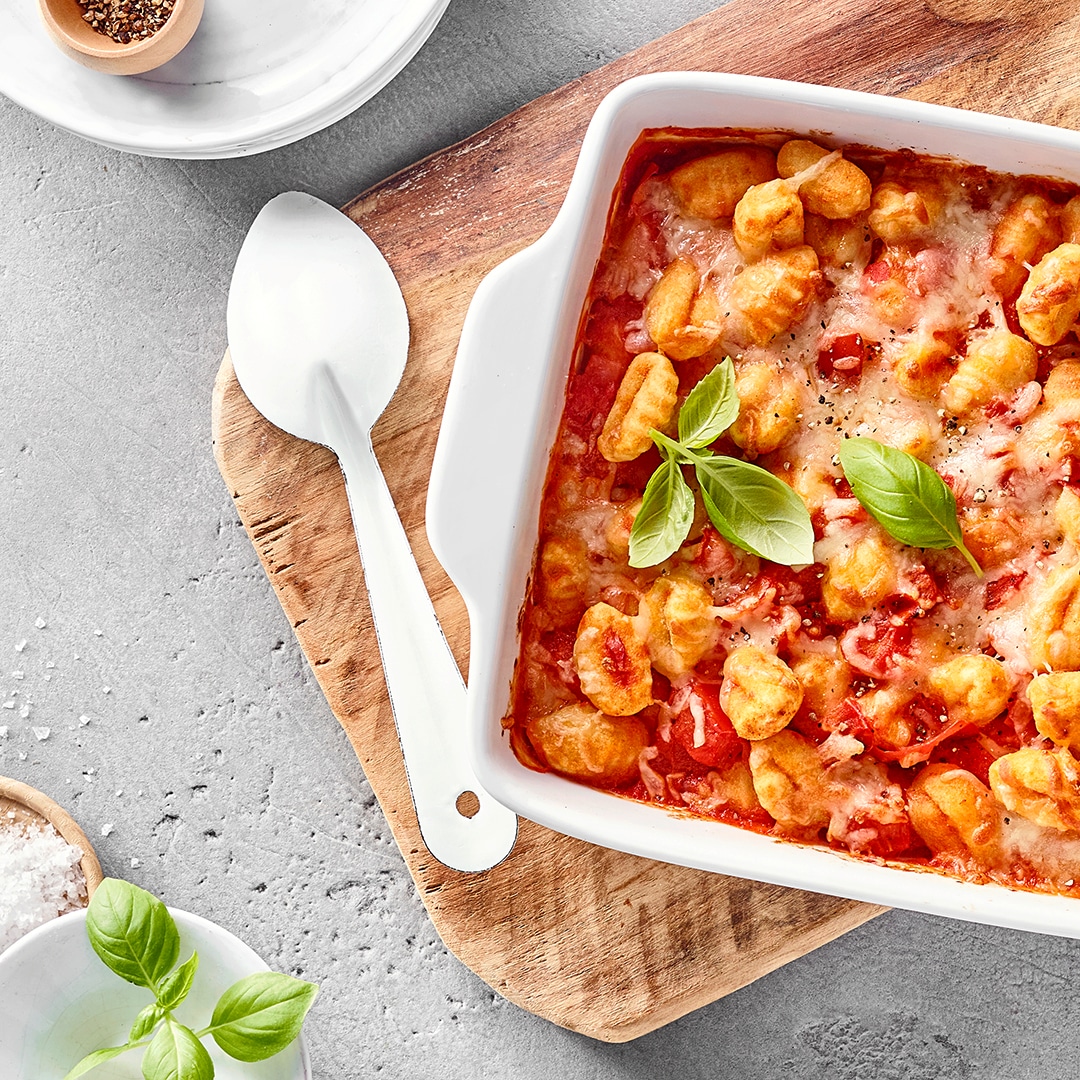 Überbackene Gnocchi mit Tomatensauce, Käse und Basilikum in einer Auflaufform auf einem Holzbrett.