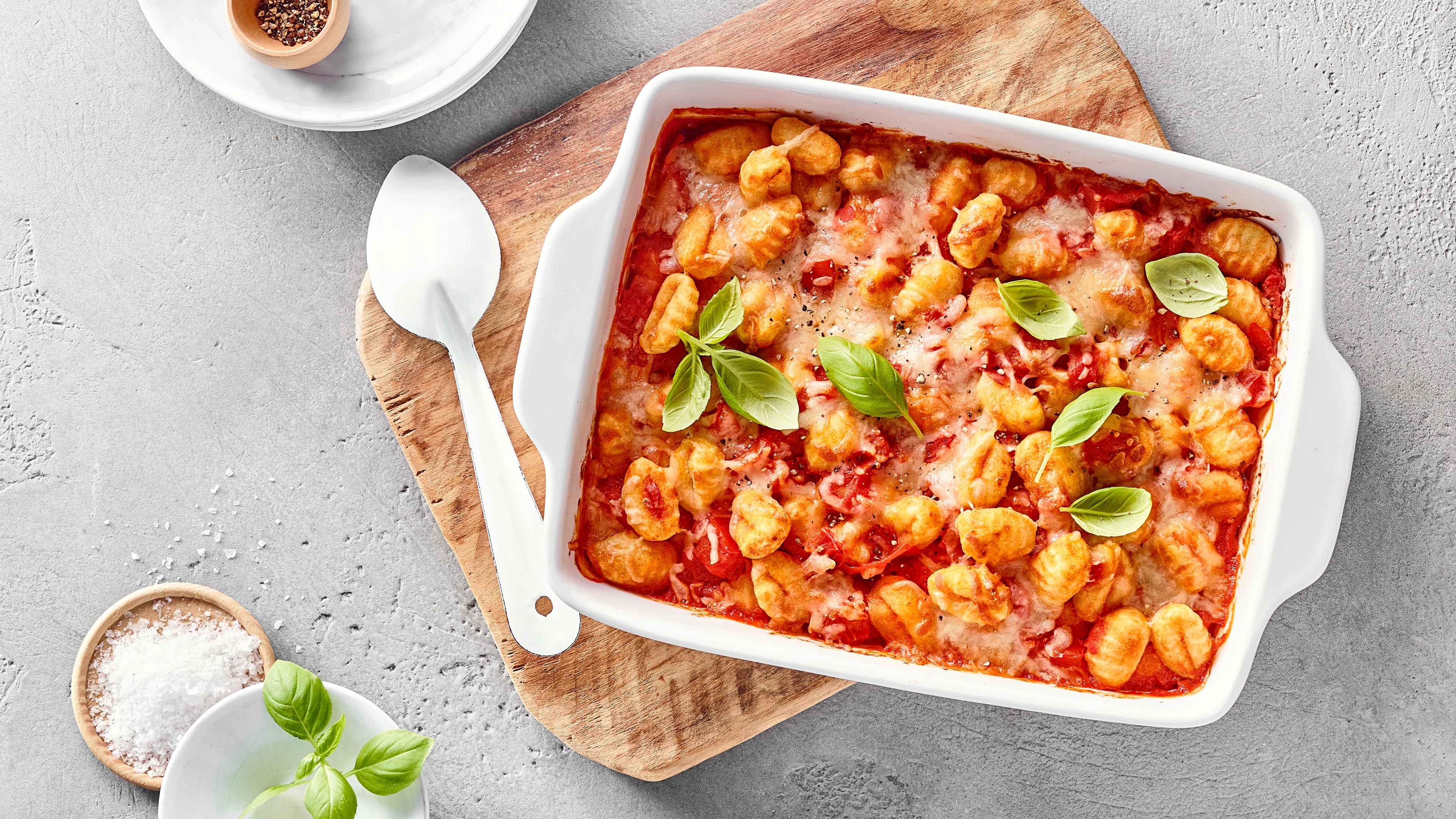 Überbackene Gnocchi mit Tomatensauce, Käse und Basilikum in einer Auflaufform auf einem Holzbrett.