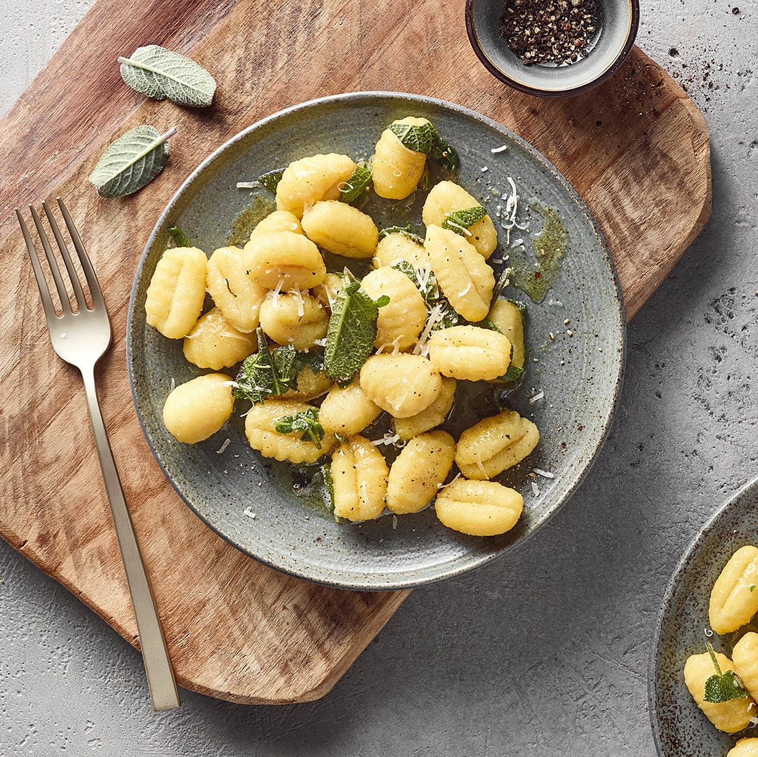 Ein Teller mit Gnocchi, darauf grüne Blätter und etwas hellem geriebenem Belag, umgeben von klarer Soße.