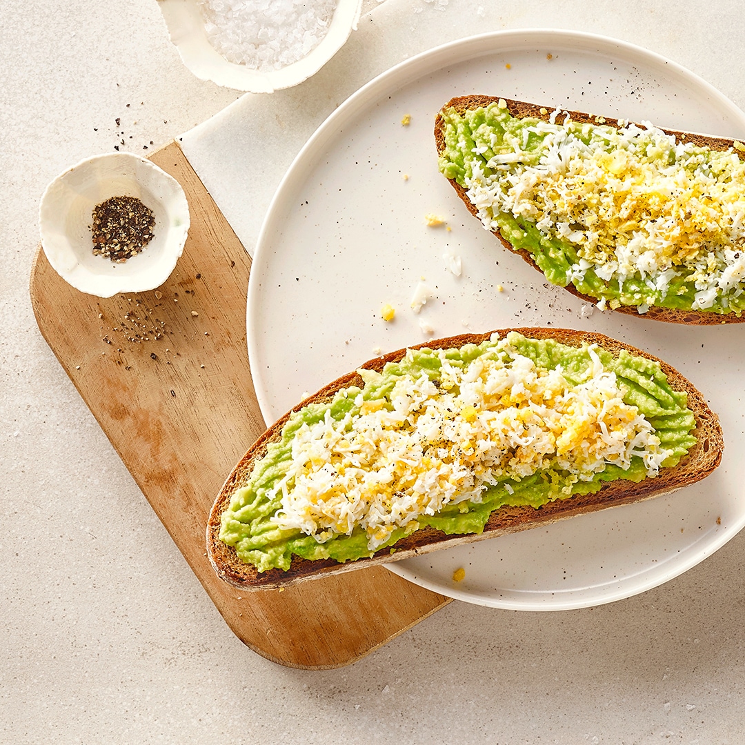 Ein Teller mit zwei Scheiben Brot mit Avocado-Aufstrich und klein geriebenem Ei, daneben Schälchen mit Salz und Pfeffer