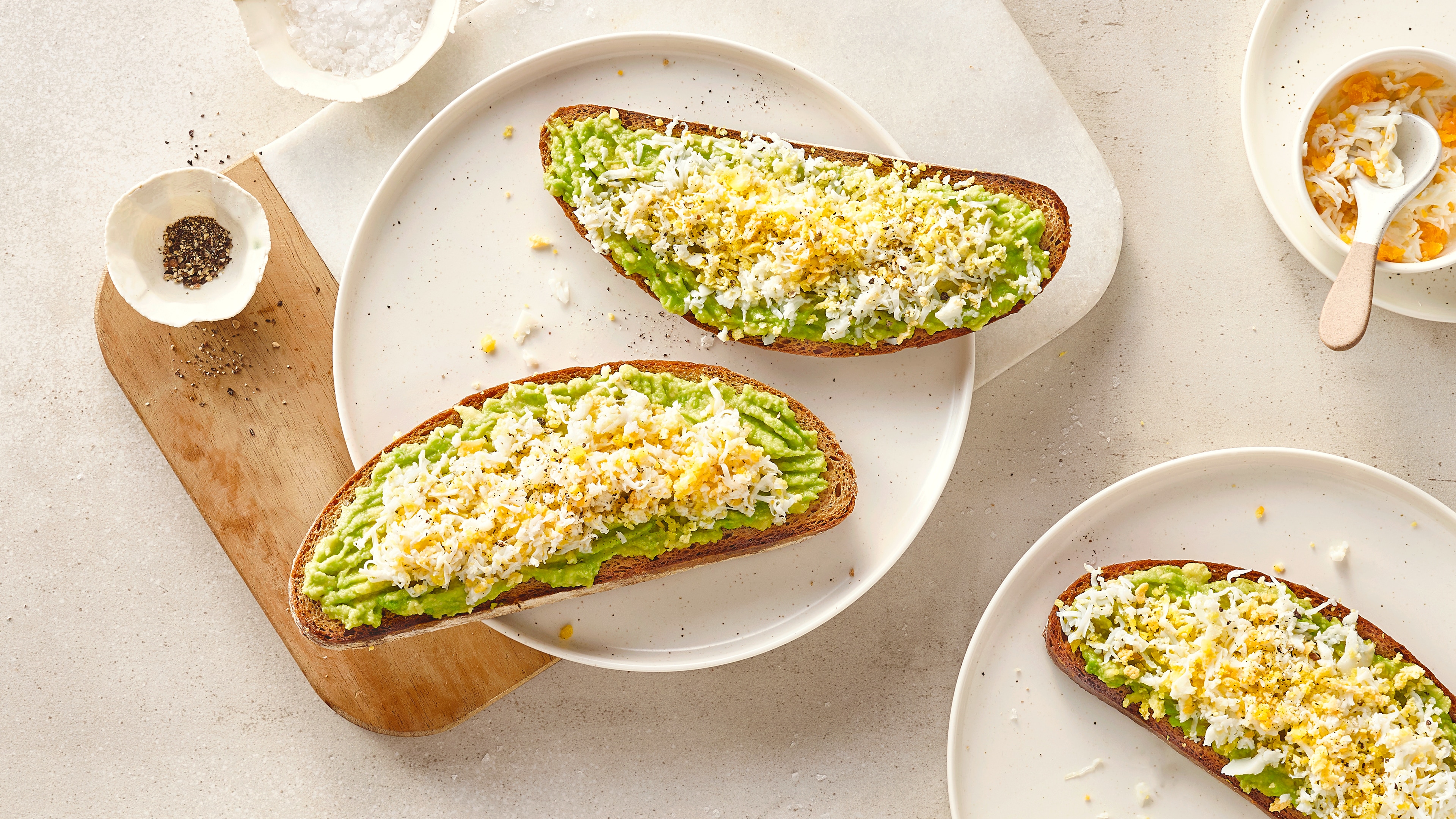 Rezept für Grated Egg Avocado Toast Ein Teller mit zwei Scheiben Brot mit Avocado-Aufstrich und klein geriebenem Ei, daneben Schälchen mit Salz und Pfeffer