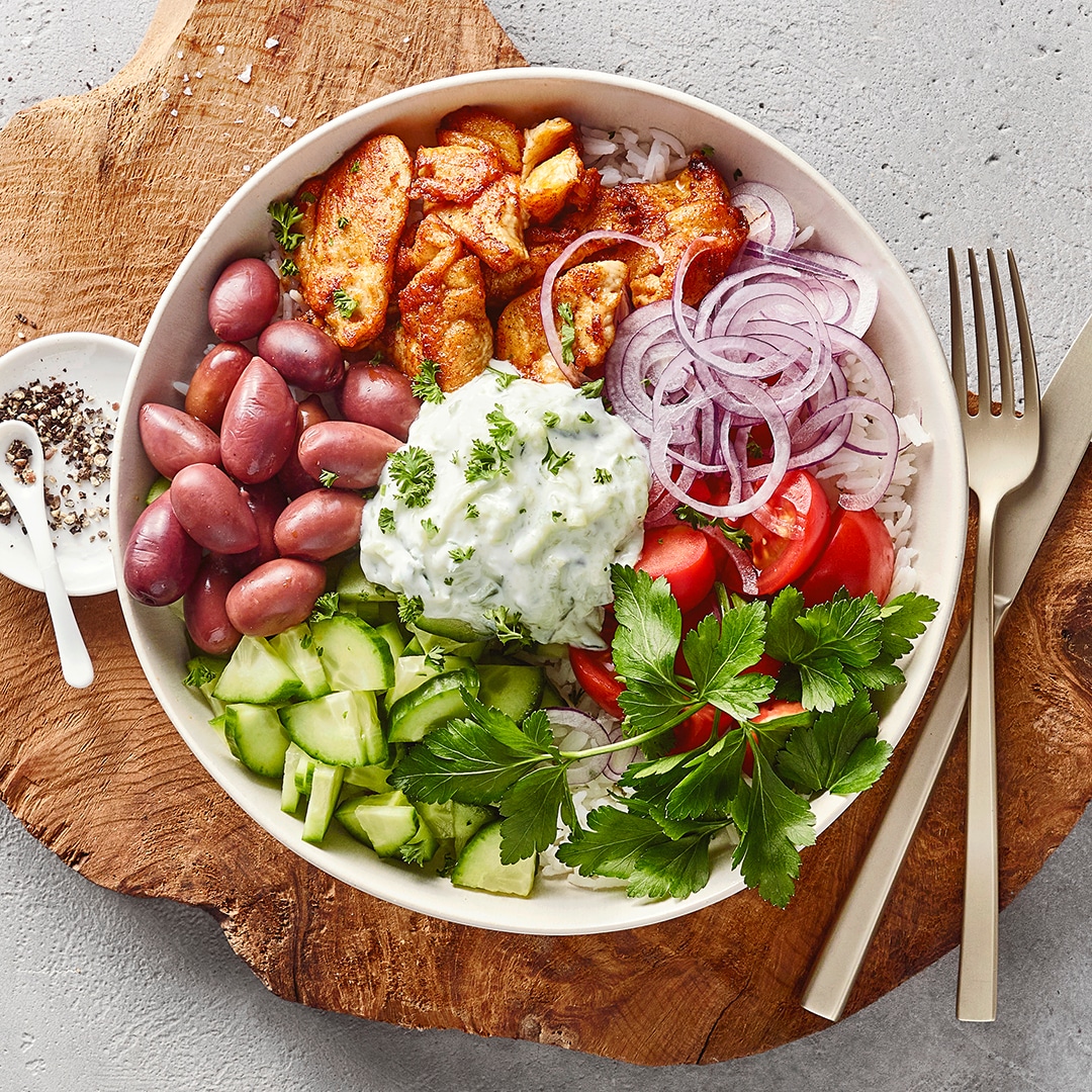 Eine Schüssel mit Hähnchen, violetten Zwiebelringen, roten Tomaten, grünen Gurken, Oliven, weißer Creme & Kräutern auf Reis.