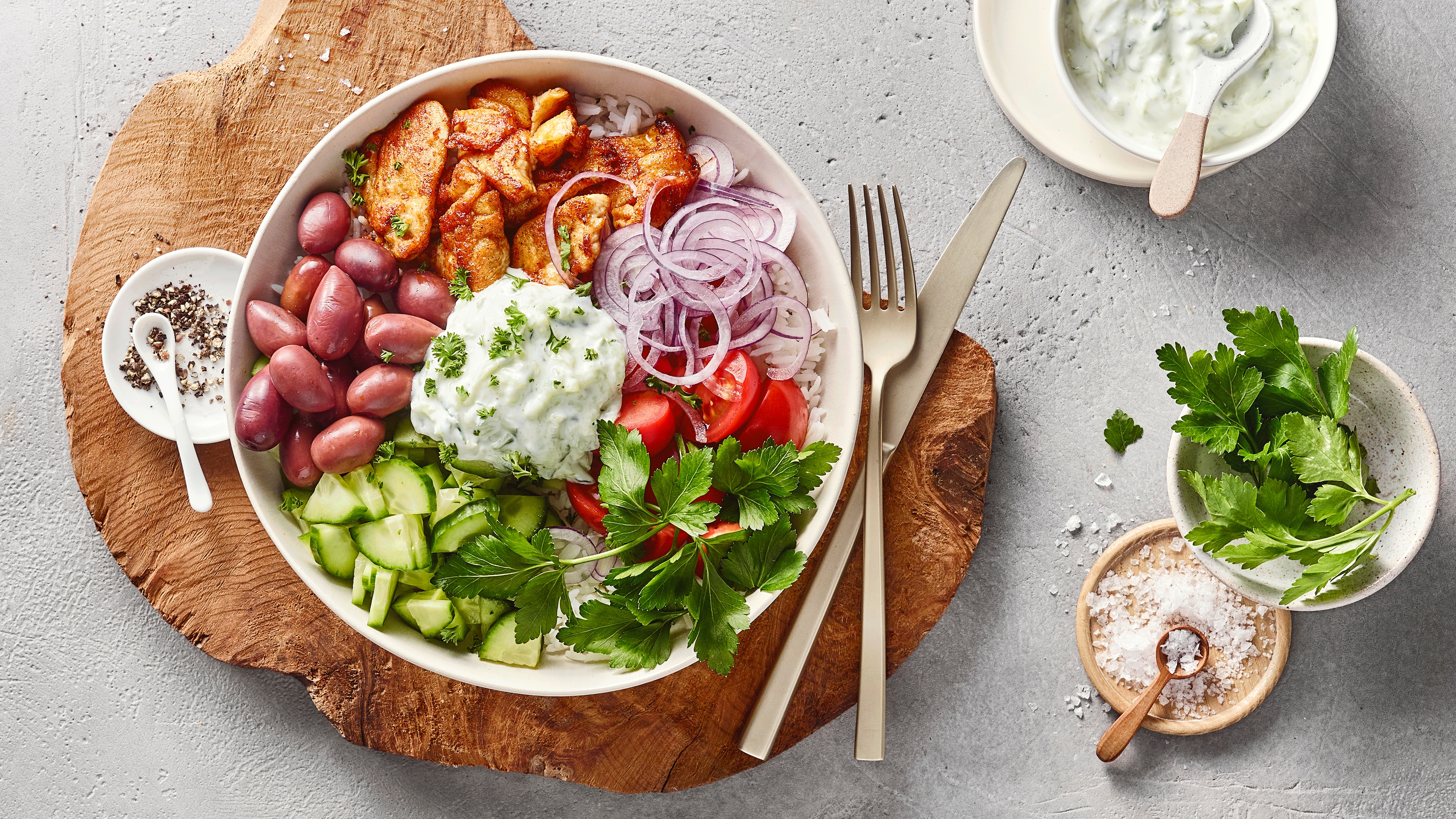 Eine Schüssel mit Hähnchen, violetten Zwiebelringen, roten Tomaten, grünen Gurken, Oliven, weißer Creme & Kräutern auf Reis.