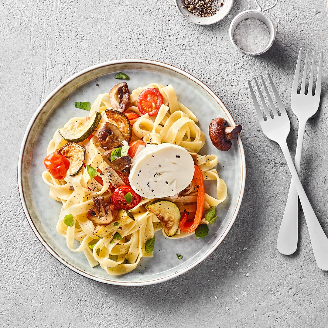 Ein Teller Bandnudeln mit gegrilltem Gemüse und Burrata, dekoriert mit frischen Kräutern.