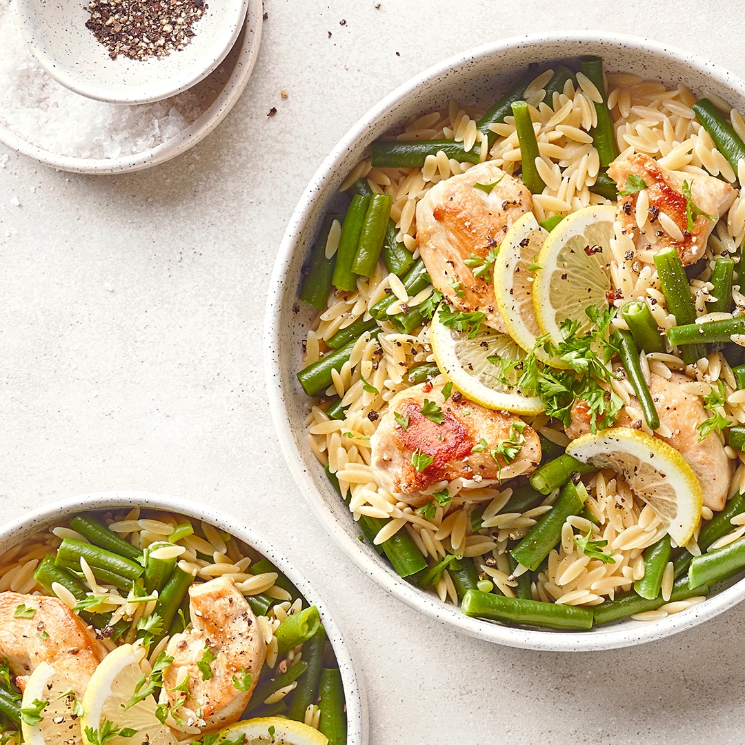 Orzo-Nudeln mit Hähnchen, grünen Bohnen und Zitronenscheiben in einer Schüssel, garniert mit Kräutern und Pfeffer.