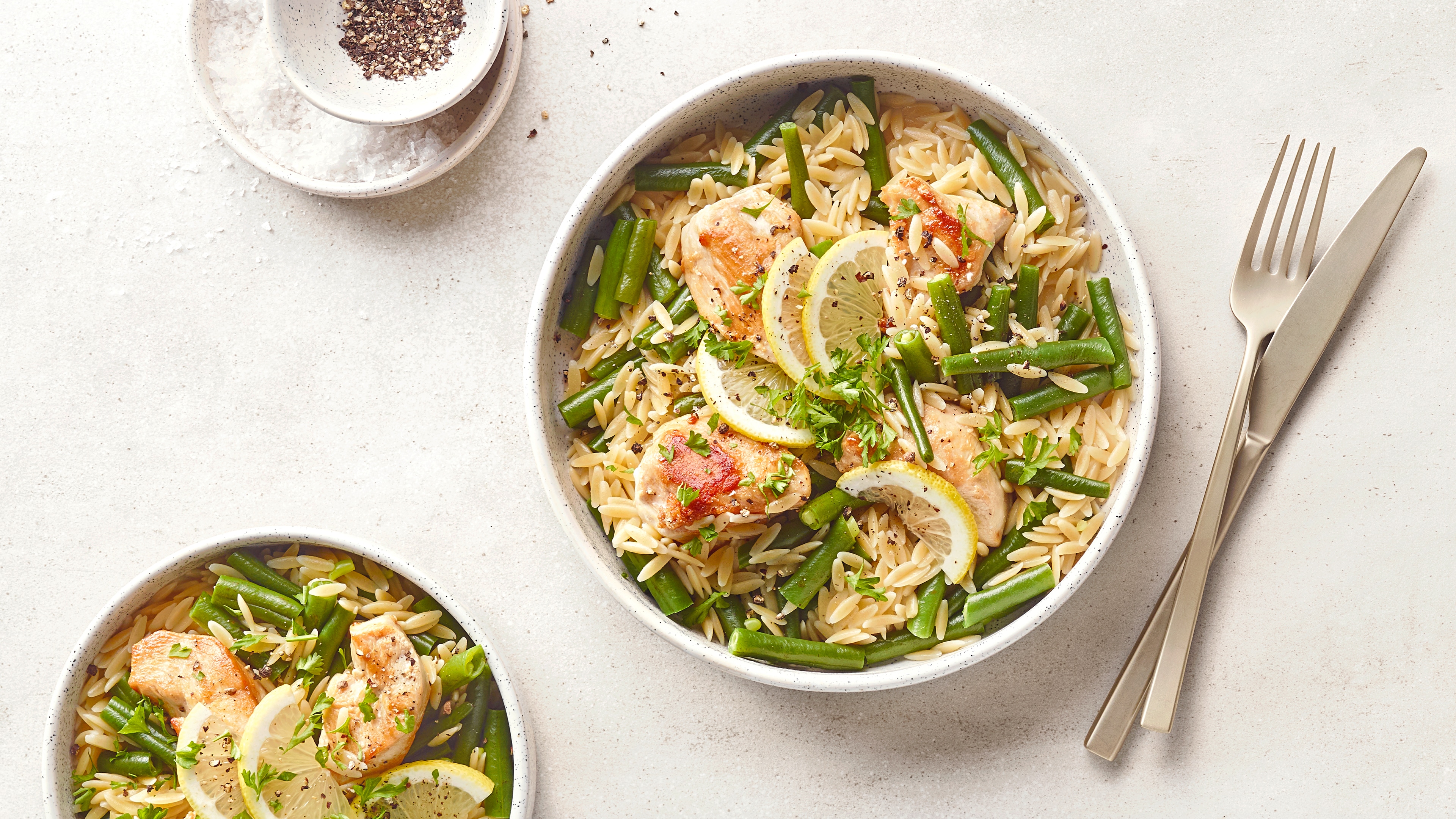 Orzo-Nudeln mit Hähnchen, grünen Bohnen und Zitronenscheiben in einer Schüssel, garniert mit Kräutern und Pfeffer.