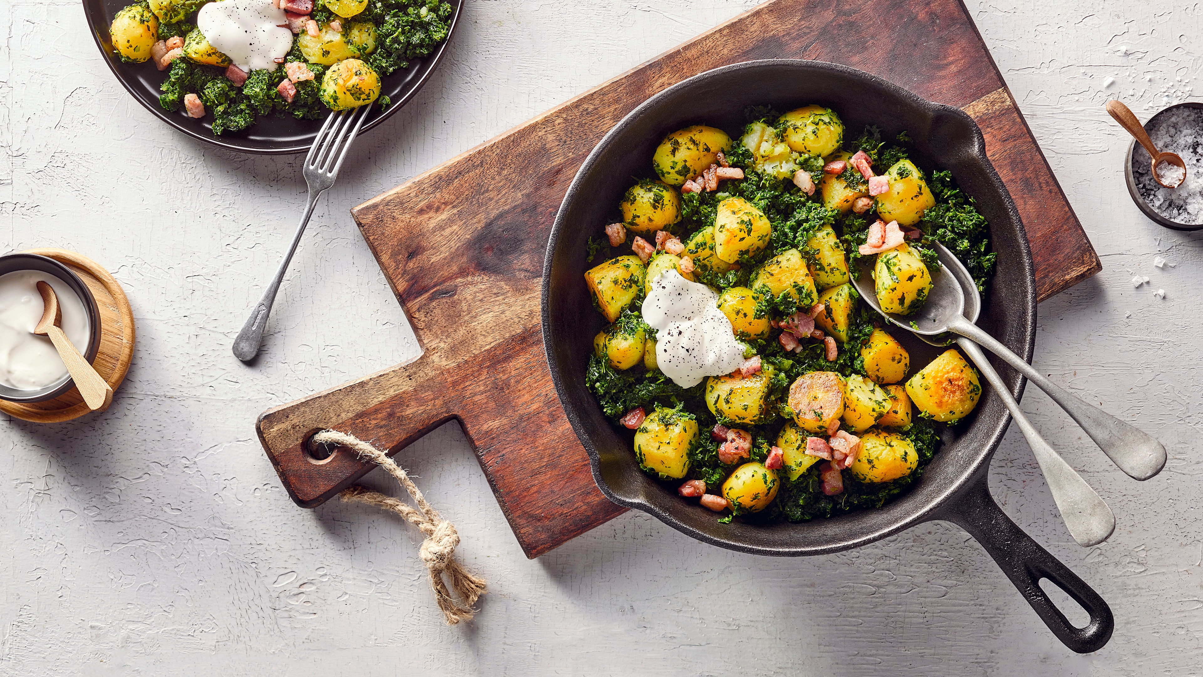 Rezept für eine Grünkohlpfanne Eine gusseiserne Pfanne mit Kartoffelstücken, Grünkohl und Speckwürfeln, getoppt mit einem Klecks weißer Soße