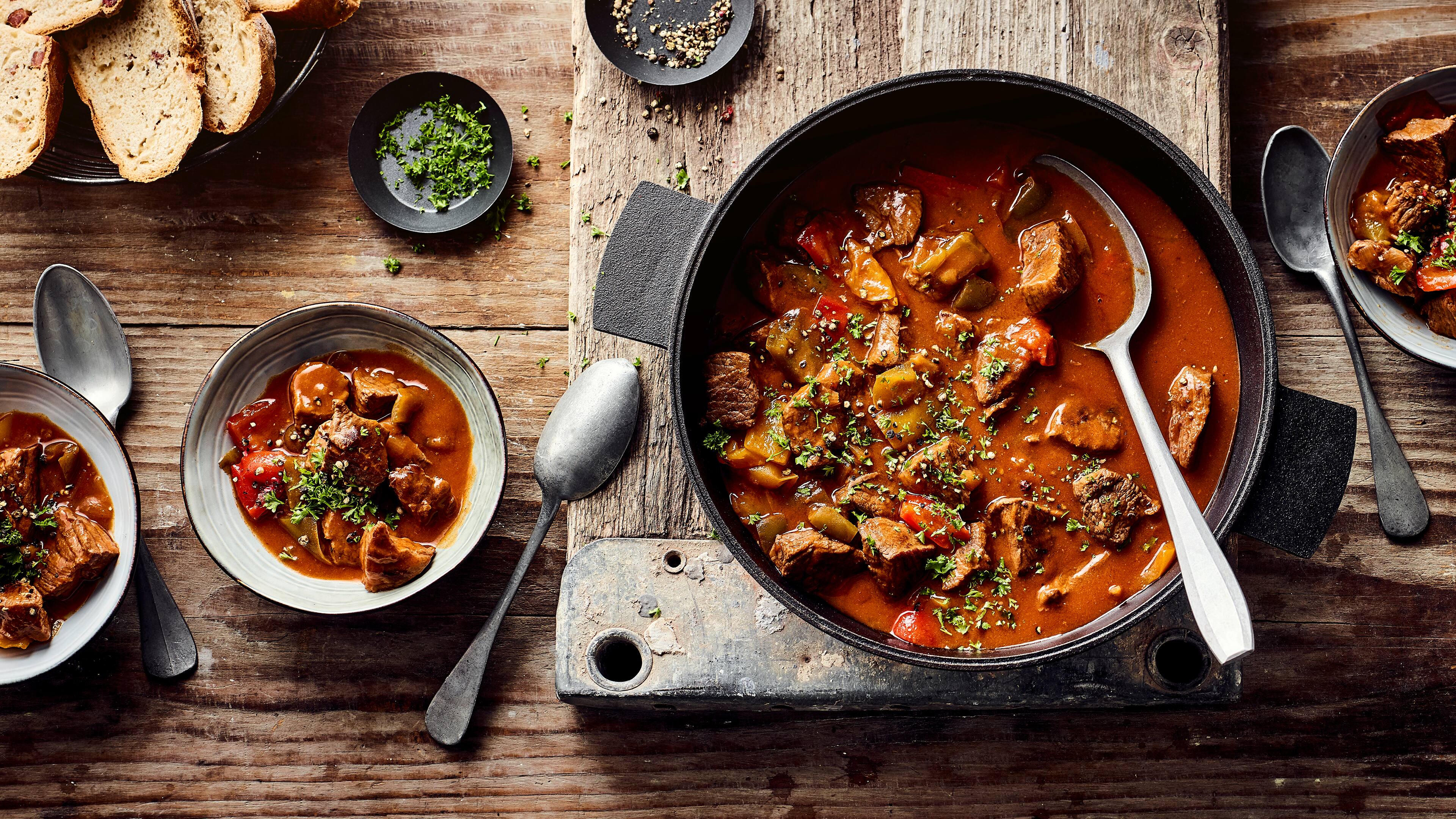 Rezept für Gulasch Dutch-Oven Ein gusseisener Topf mit Fleisch und Paprika in rotbrauner Soße, getoppt mit frischer, gehackter Petersilie