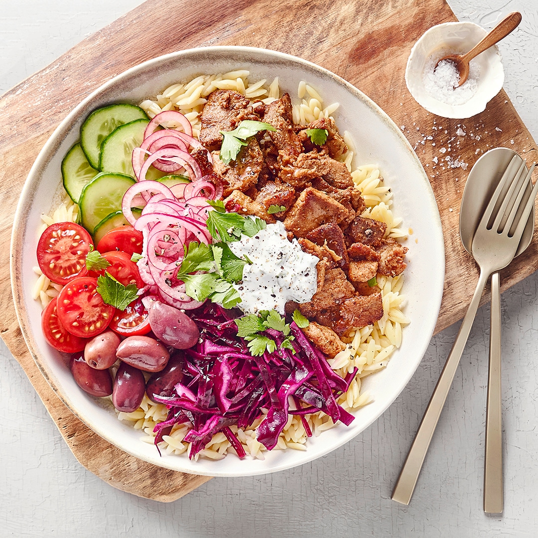 Tiefer Teller auf Brett gefüllt mit Reis, Gurken, Tomaten, Fleisch, Zwiebeln, rotem Krautsalat, Oliven und Tzaziki.