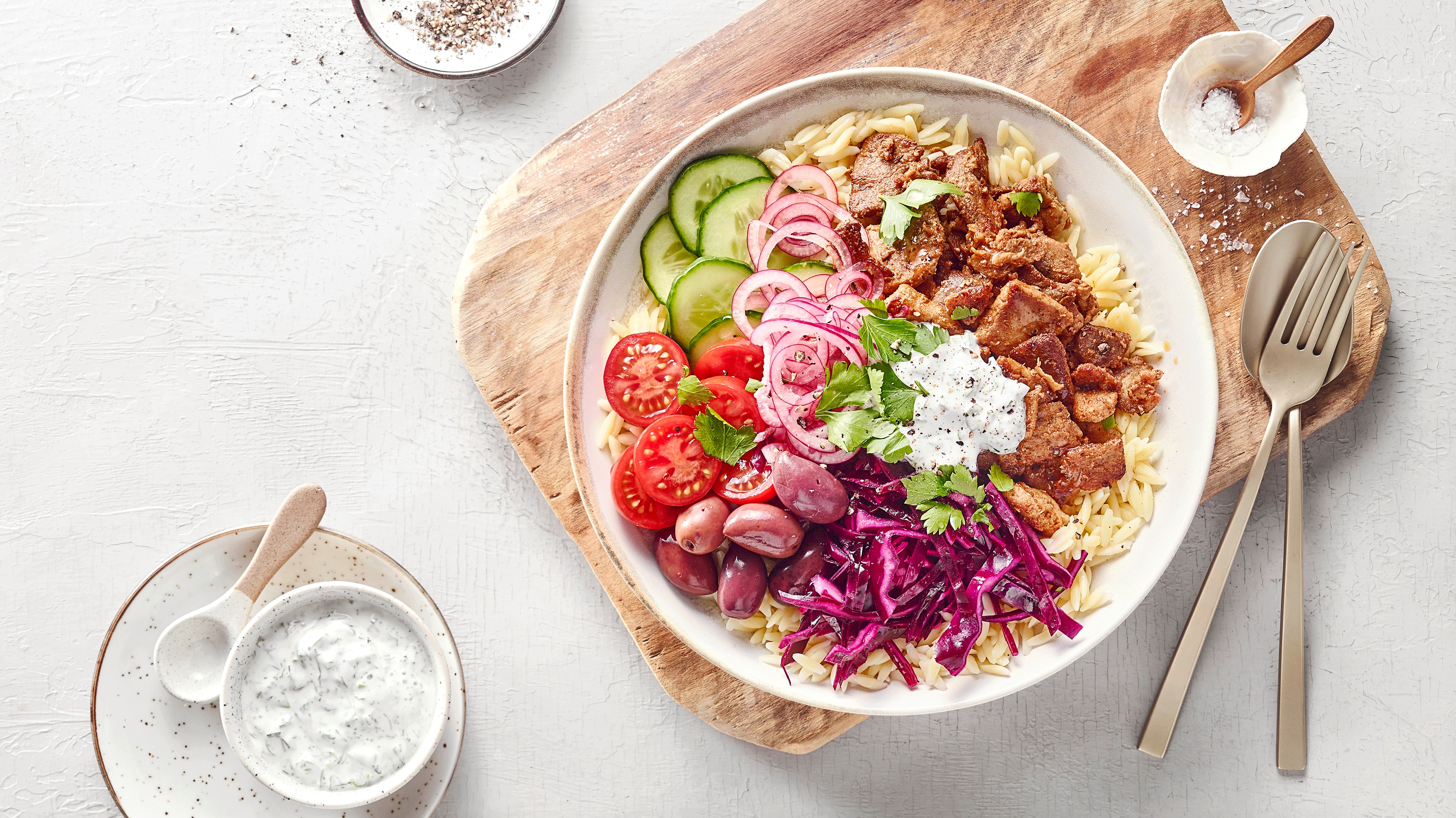 Tiefer Teller auf Brett gefüllt mit Reis, Gurken, Tomaten, Fleisch, Zwiebeln, rotem Krautsalat und Oliven. Daneben ein Schälchen mit Tzaziki.