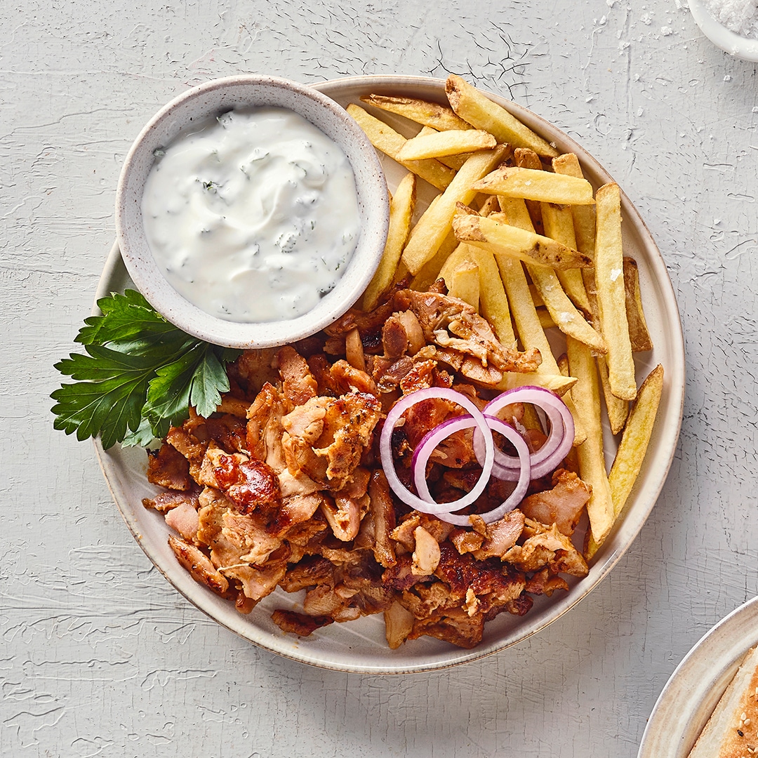 Ein Teller mit goldgelben Pommes, gebratenen braunen Fleischstücken, roten Zwiebelringen, grünen Kräutern und einer Schale weißer Sauce.