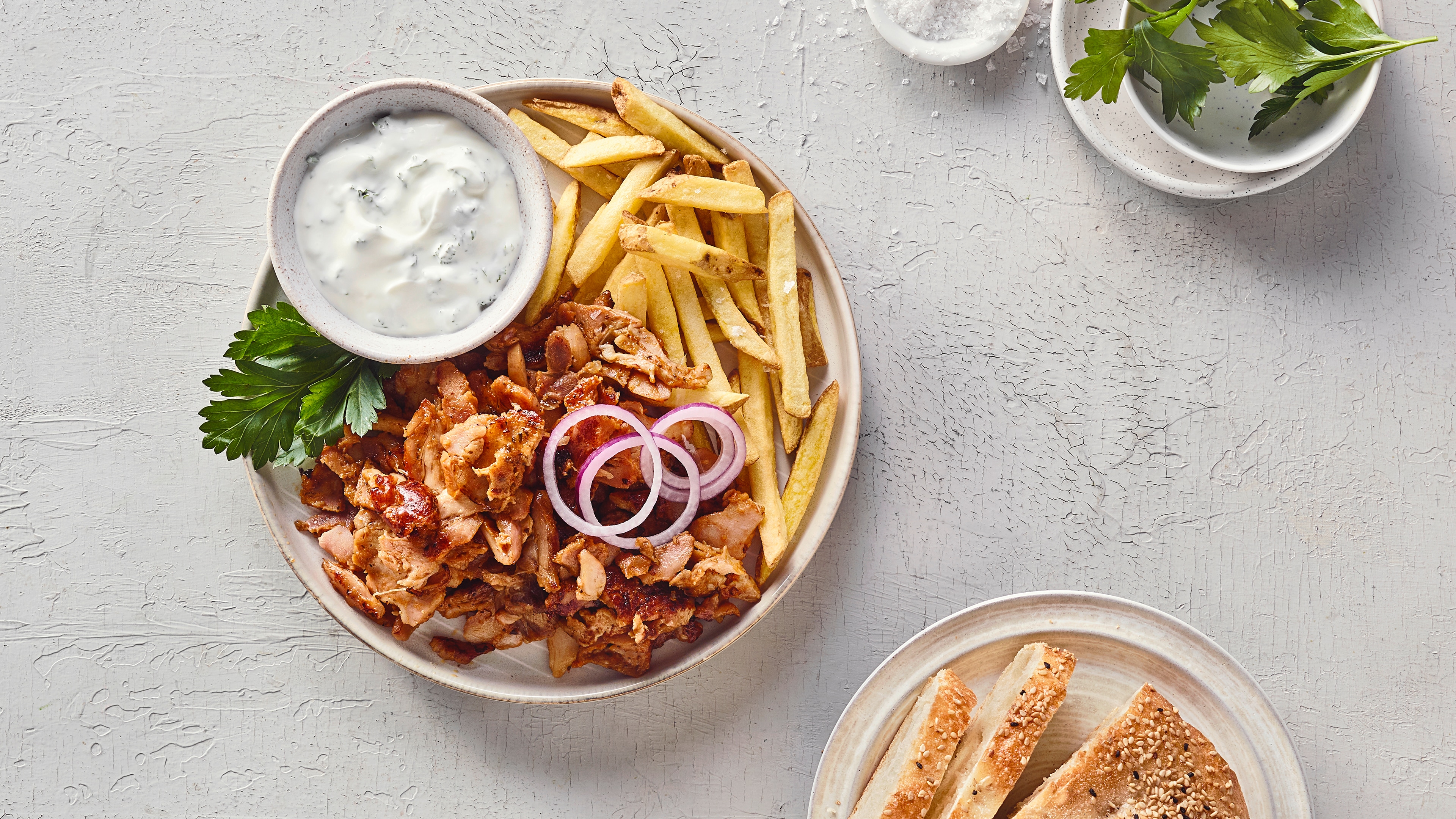 Ein Teller mit goldgelben Pommes, gebratenen braunen Fleischstücken, roten Zwiebelringen, grünen Kräutern und einer Schale weißer Sauce.