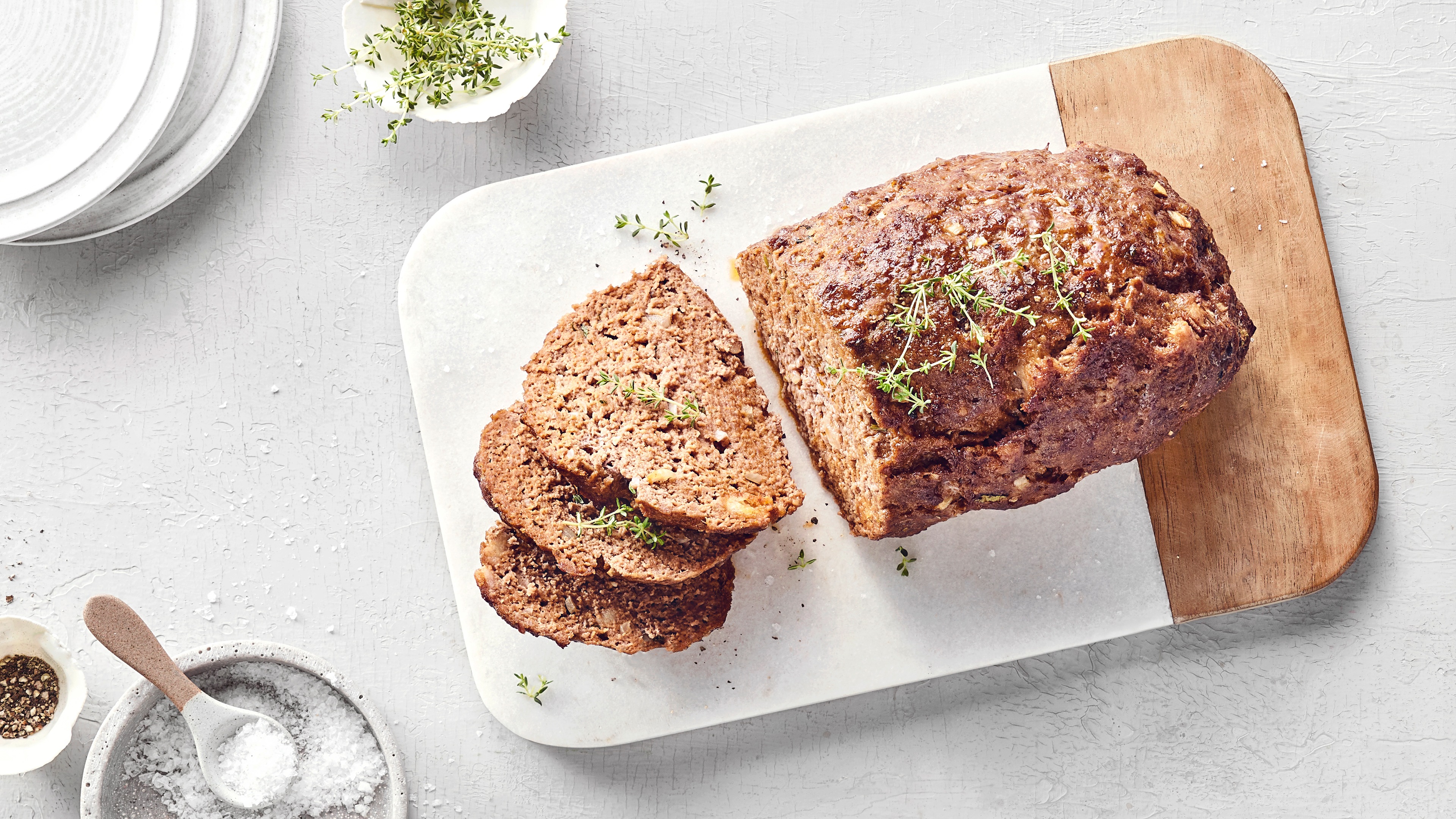 Hackbraten auf einem Schneidebrett, teilweise in Scheiben geschnitten, mit Thymian bestreut. Daneben Schälchen