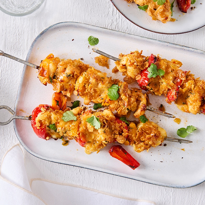 Drei Spieße mit cornflakes‑umhüllten Hähnchenstücken und Paprika. Teilweise zu sehen sind oben links ein Glas Wasser und oben rechts ein Teller mit weiteren Hähnchenspießen.