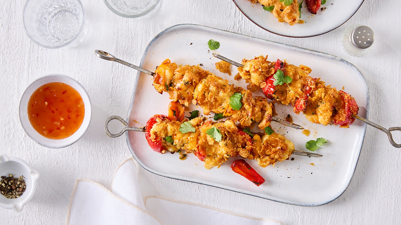 Drei Spieße mit cornflakes‑umhüllten Hähnchenstücken und Paprika. Links daneben steht eine kleine Schale mit süß-saurem Dip. Oben links stehen zwei Gläser Wasser. Oben rechts ist ein Teller mit weiteren Hähnchenspießen teilweise zu sehen.