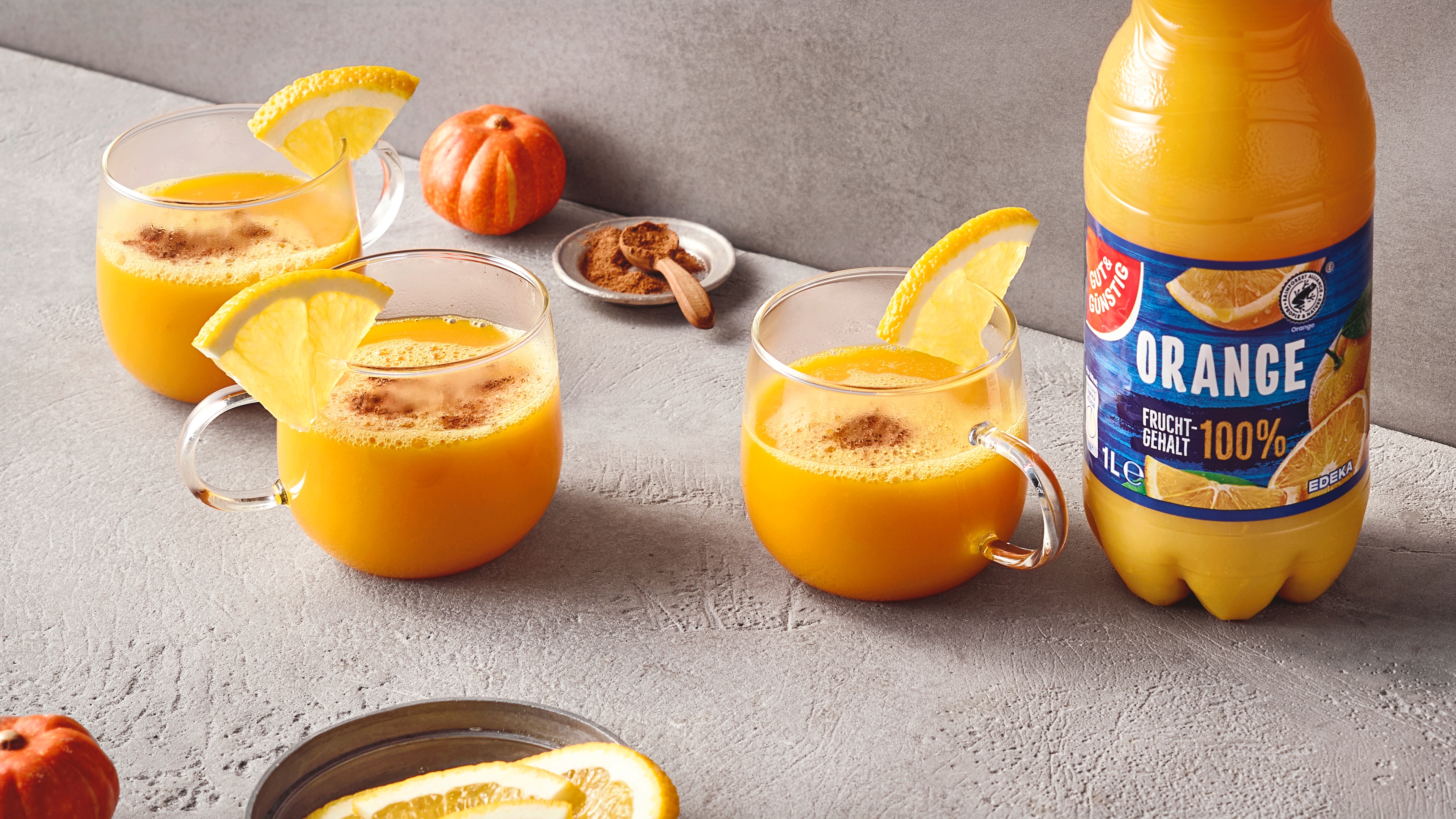 Drei Gläser mit heißem Orangensaft und Zitronenscheiben auf einem Tisch, daneben eine Flasche GUT&GÜNSTIG Orangensaft.