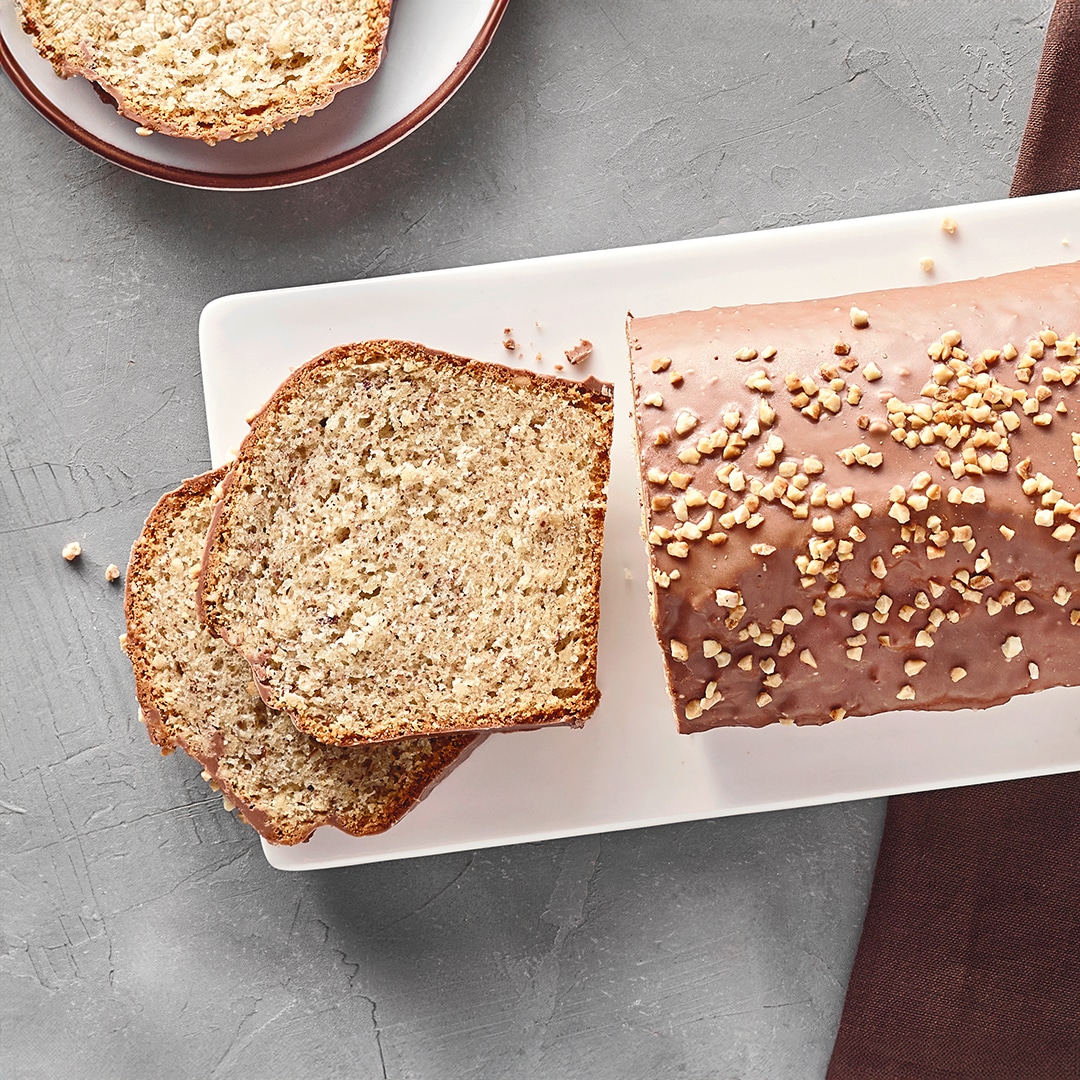 Ein Teller mit einem Kastenkuchen, überzogen mit Schokolade, garniert mit gehackten Nüssen, zwei abgeschnittene Stücke daneben