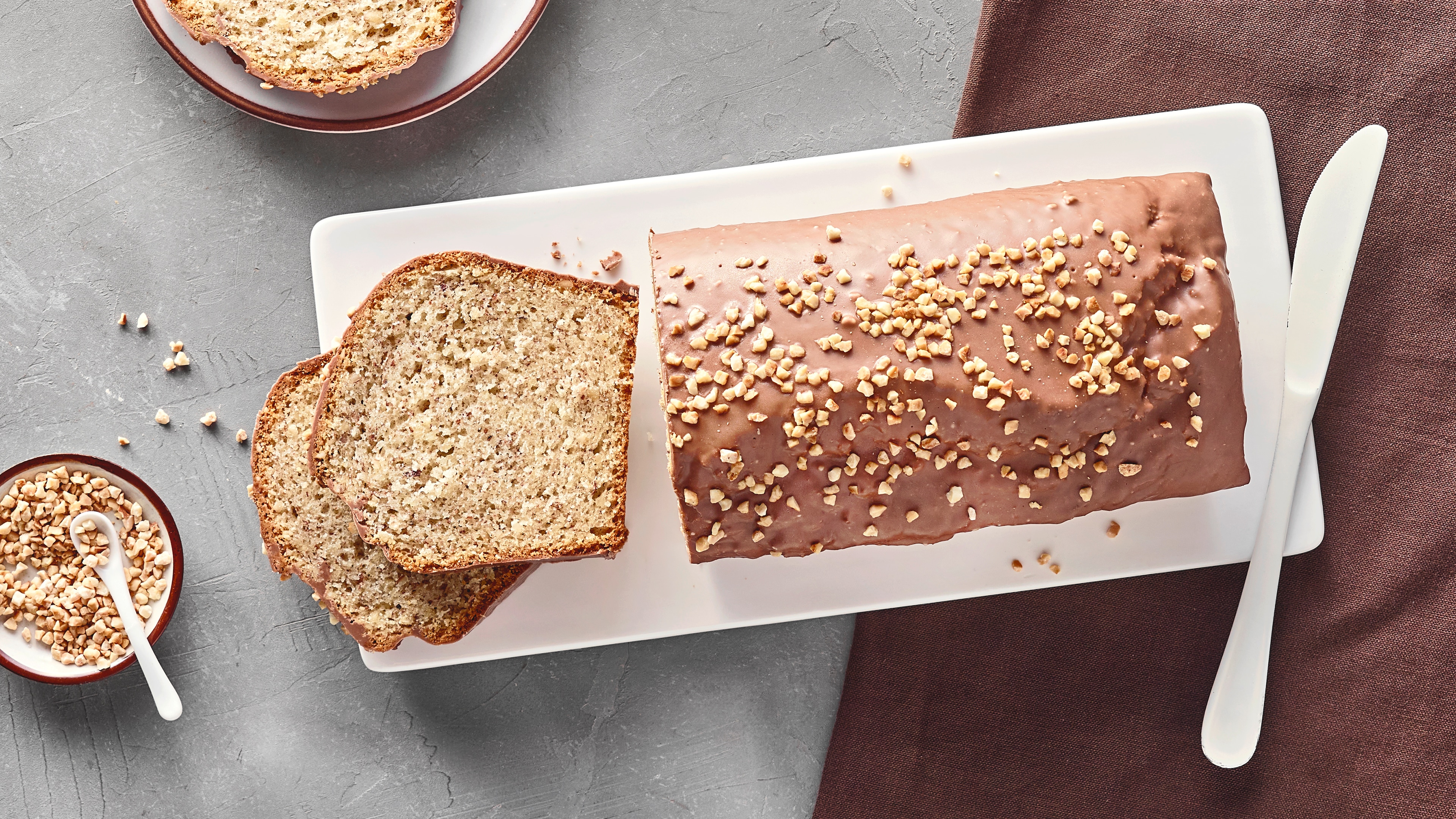 Ein Teller mit einem Kastenkuchen, überzogen mit Schokolade, garniert mit gehackten Nüssen, zwei abgeschnittene Stücke daneben