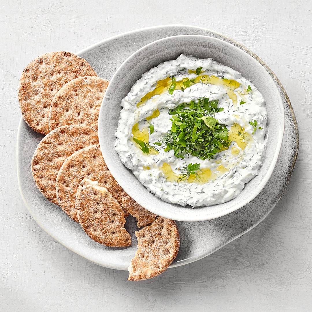 Schälchem mit weißem, leicht stückigem Dip, darauf Kräuter und Öl. Daneben Cracker