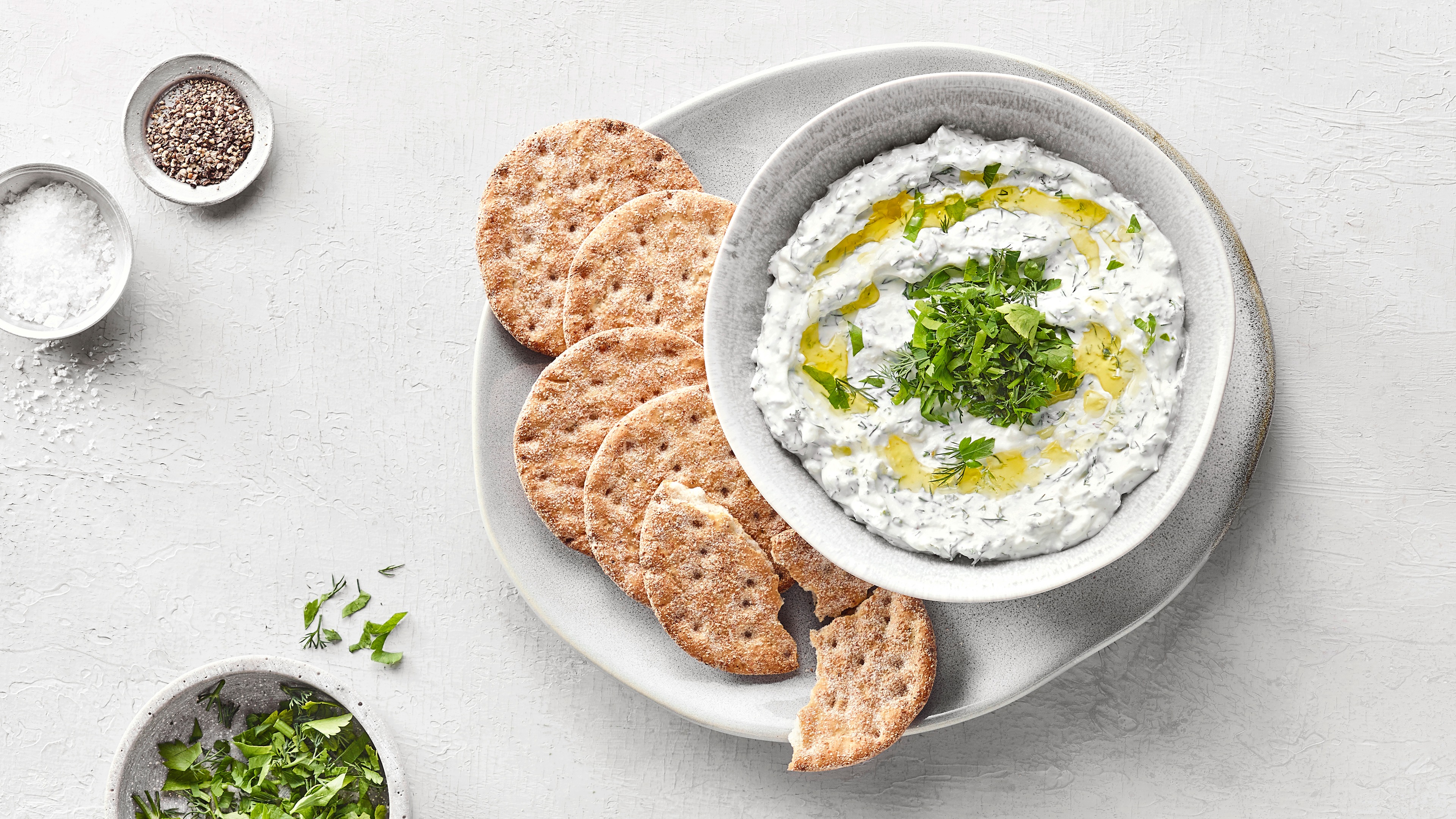 Schälchem mit weißem, leicht stückigem Dip, darauf Kräuter und Öl. Daneben Cracker auf einem Teller und Schälchen mit Salz, Pfeffer und Kräutern
