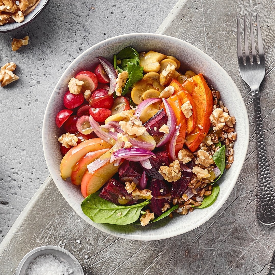 Eine Schüssel mit roten Trauben, Apfelscheiben, Kürbisstücken, roten Zwiebeln, grünen Blättern und Walnüssen.